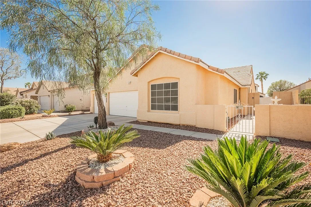 Front exterior view of a single-story Las Vegas home featuring desert landscaping with decorative rock, mature trees, and low-maintenance plants. The home has a light stucco exterior, a two-car garage, a large front window, and a gated courtyard entr