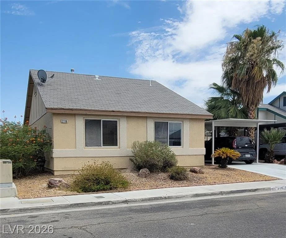Front exterior view of a single-story beige home with a steep gabled roof, desert landscaping, and a covered carport. The property features mature palm trees, low-maintenance rock landscaping with shrubs, and a concrete driveway in a quiet residentia