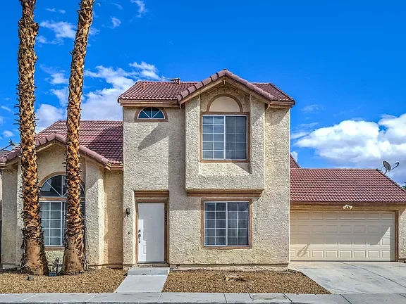 Front exterior view of a two-story Las Vegas home featuring a light stucco façade, tiled roof, and attached two-car garage. The property includes low-maintenance desert landscaping, mature palm trees, and large windows that provide natural light. Set
