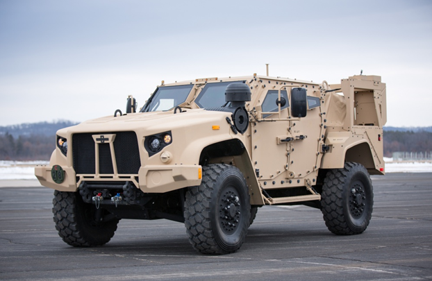 A tan military armored vehicle parked on an open tarmac with a cloudy sky and snow in the background.