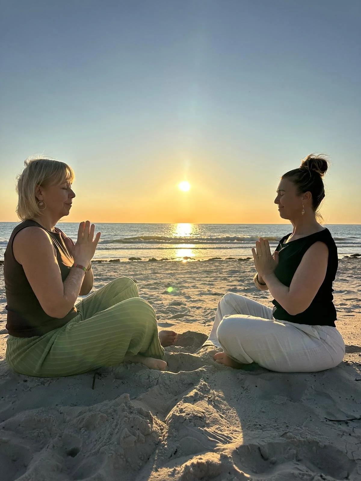 Two women sitting cross-legged on the beach facing each other with hands pressed together in a meditation pose during sunset.