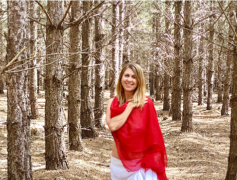 A woman standing in a forest with tall trees, wearing a red shawl and white pants, smiling at the camera.