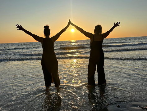 Two women standing in the ocean at sunset, facing away, with arms raised and touching hands, creating a silhouette against the sun.