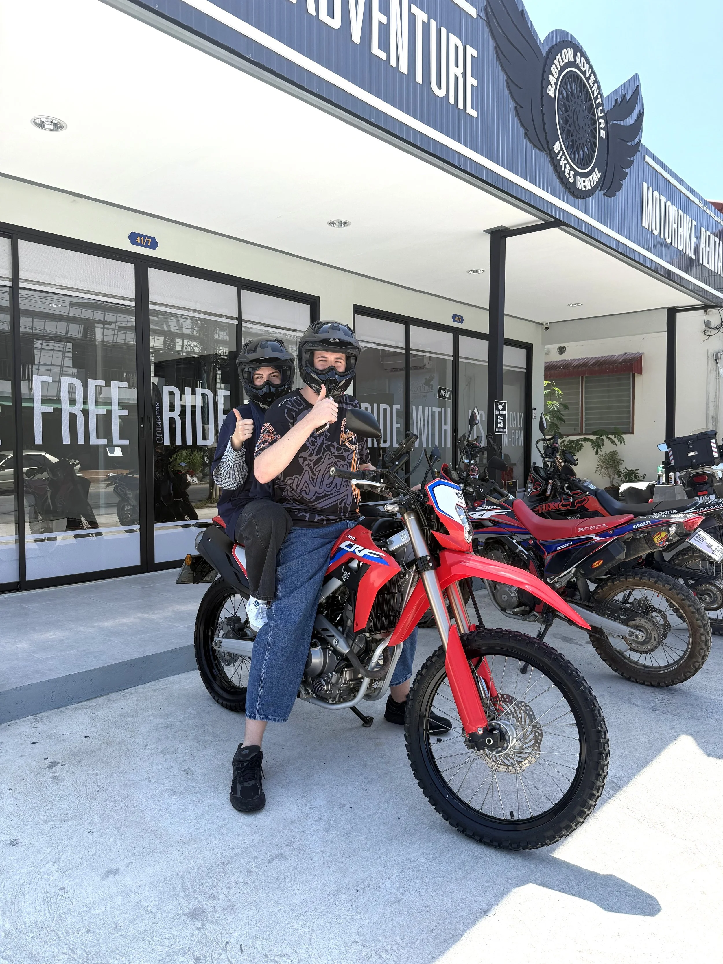 Two individuals wearing helmets and face masks on a red electric dirt bike outside a motorcycle rental shop. One person is sitting on the bike, giving a thumbs-up, and the other is standing behind them. There are additional motorcycles parked beside 