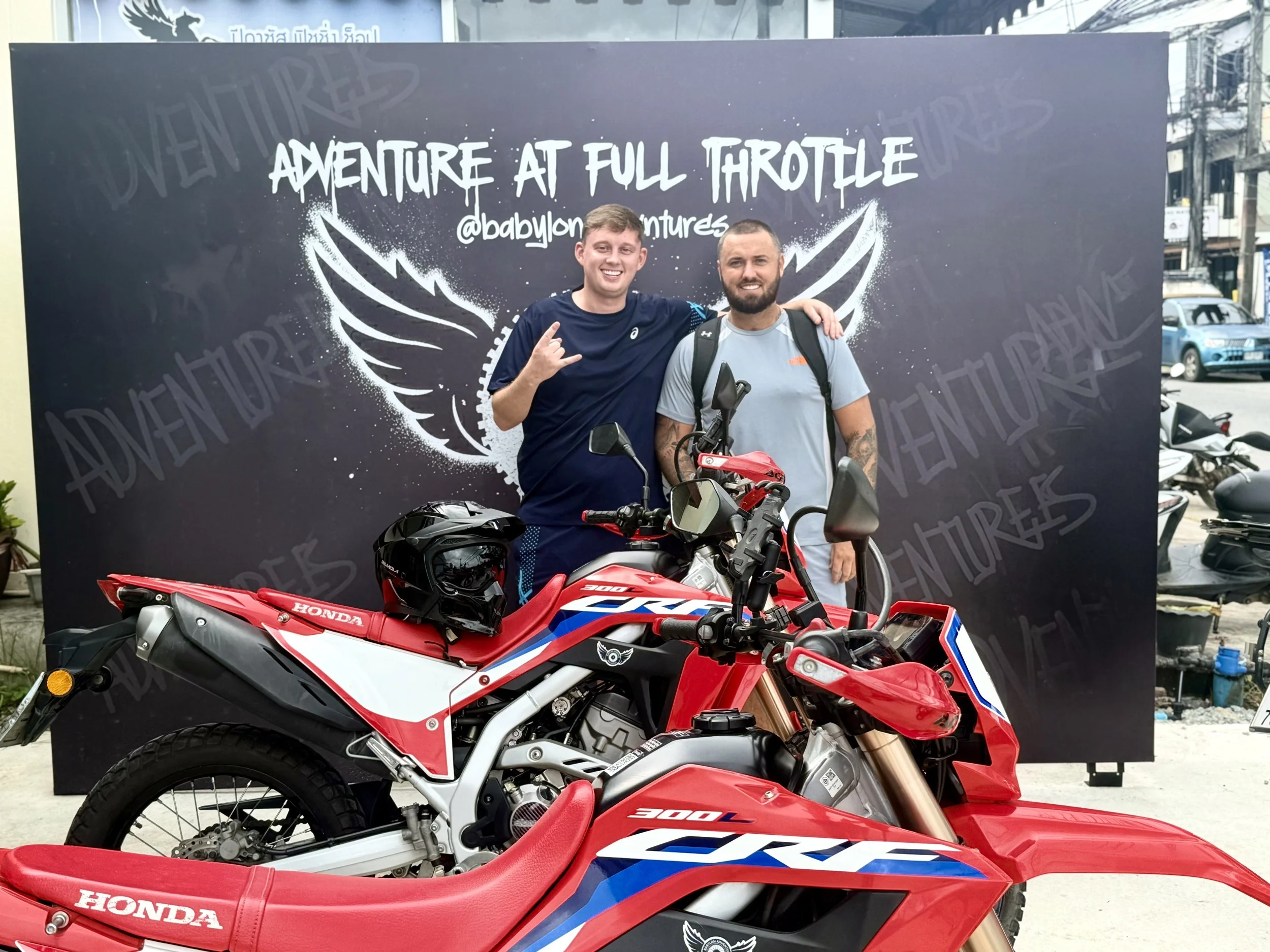 Two men standing in front of a backdrop with the words 'Adventure at Full Throttle' and '@babylonadventures', smiling with one arm around each other's shoulder. There are two red and white Honda CRF motorcycles in front of them, one with a black helm