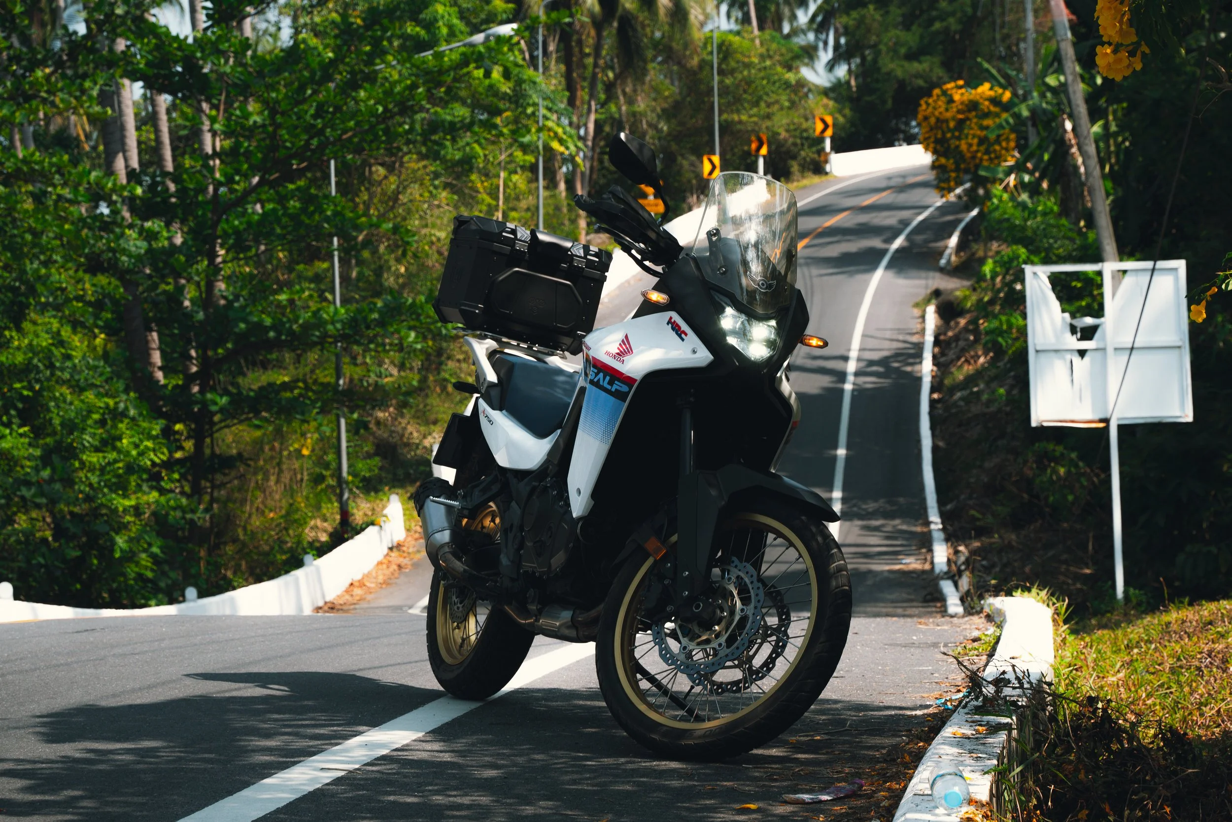 A white and black Honda adventure motorcycle parked on the side of a winding mountain road surrounded by green trees and foliage.