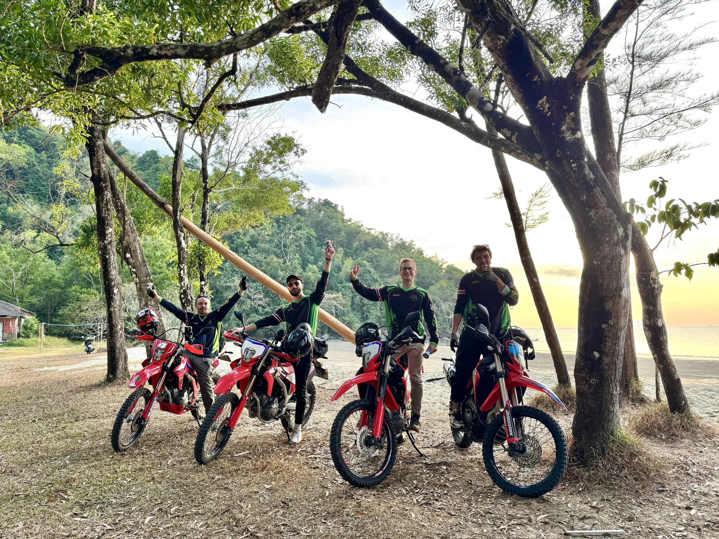 Four men in black and green motocross gear with red motocross bikes standing on a dirt clearing near the ocean at sunset, with trees and hills in the background.