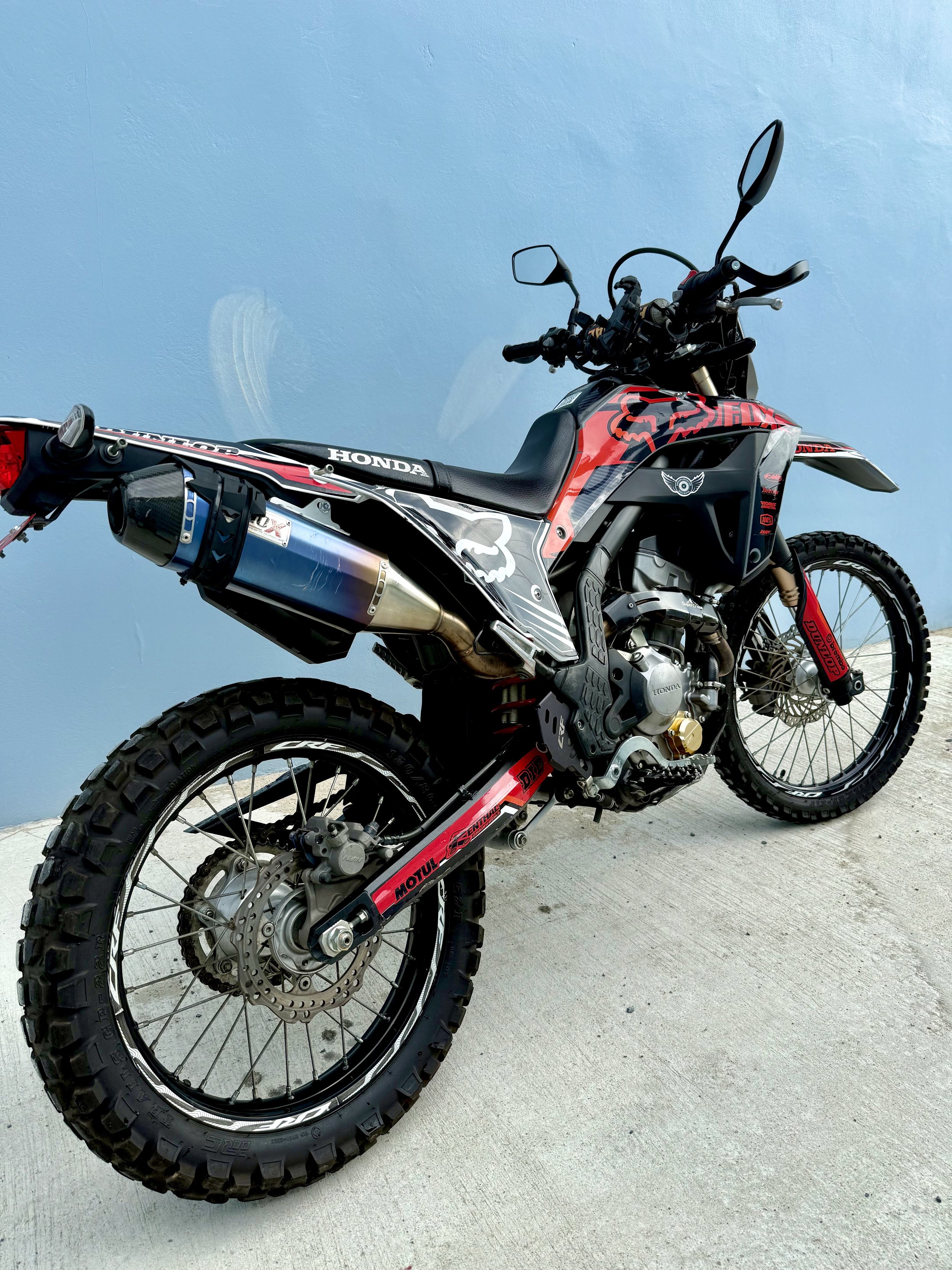 A black and red off-road Honda dirt bike with rugged tires, sitting against a light blue wall.