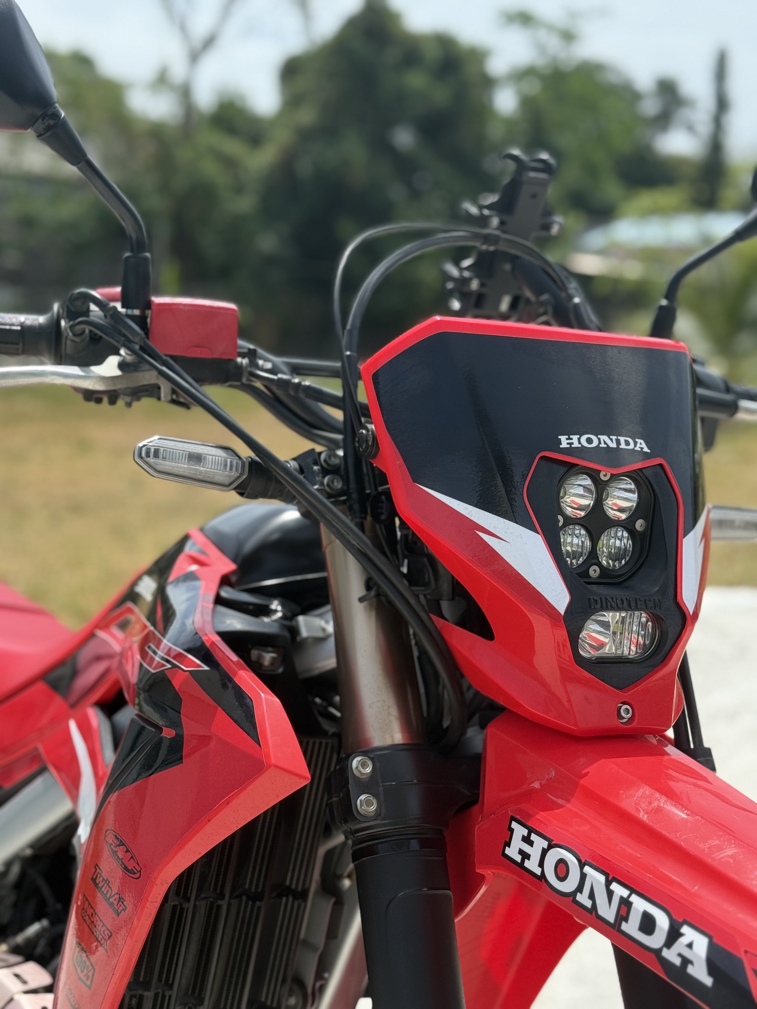 Close-up of a red Honda dirt bike with a black and white headlight cover, handlebars, and front suspension visible. The background is blurred with trees and sky.