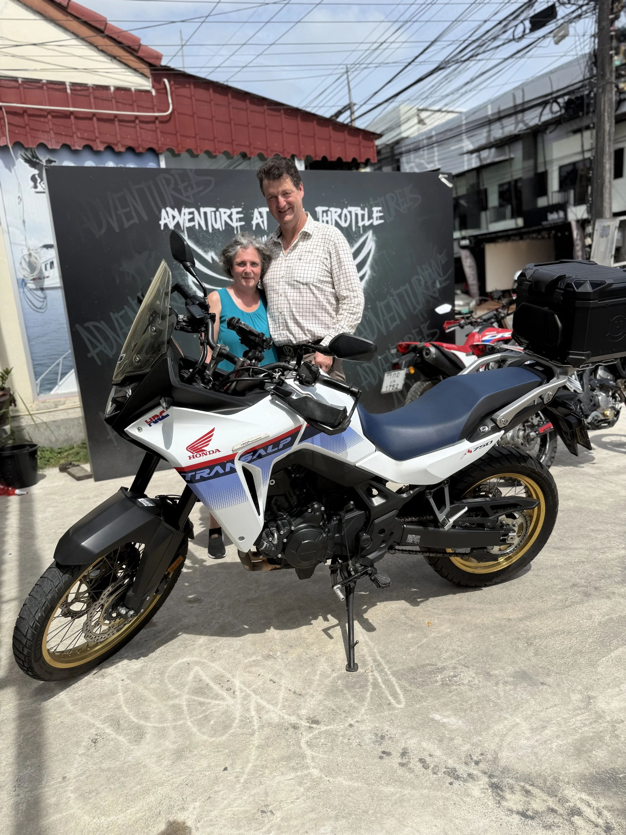 A man and a woman standing behind a Honda off-road motorcycle, smiling for the photo at an outdoor event with a black backdrop that reads 'Adventure at Full Throttle'.