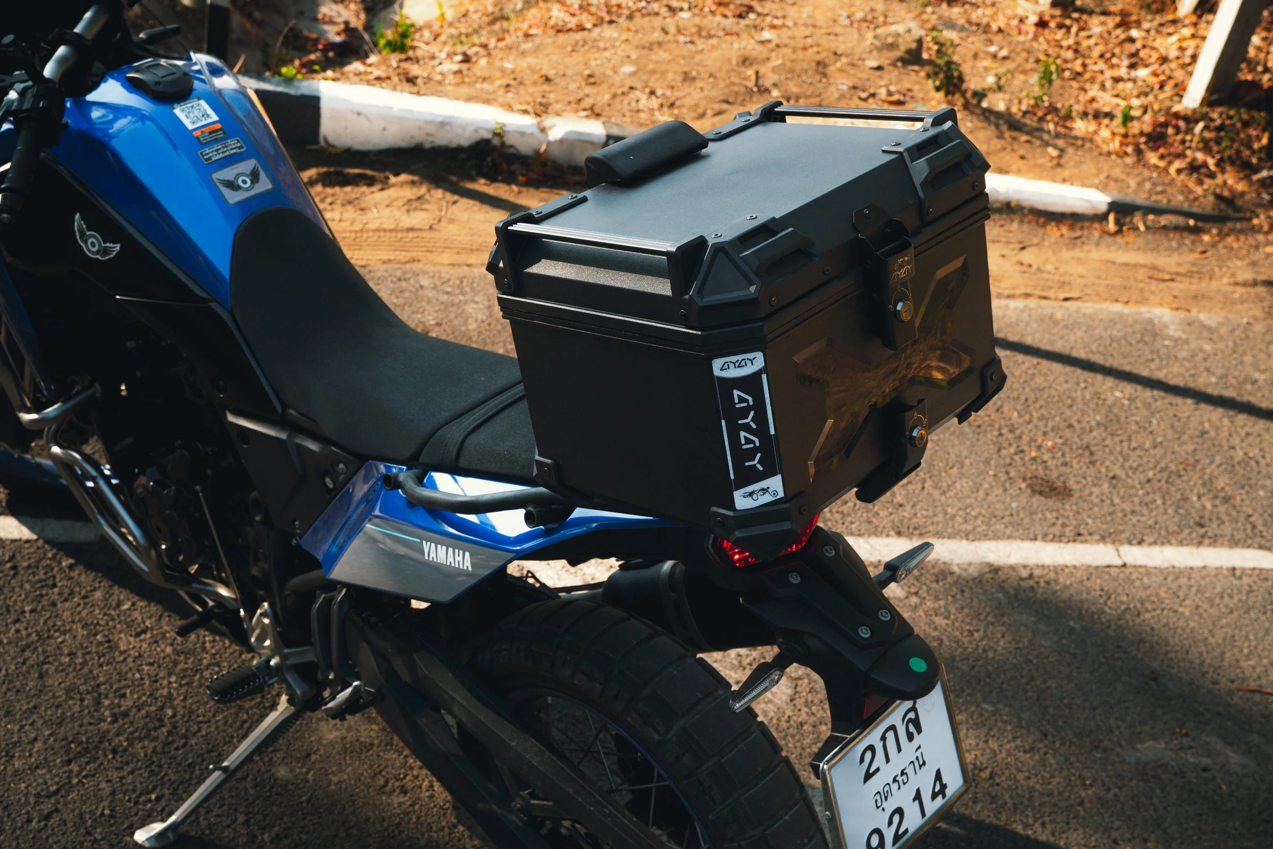 A blue Yamaha motorcycle with a black storage box mounted on the rear, parked on a paved area with a dirt and leaf background.