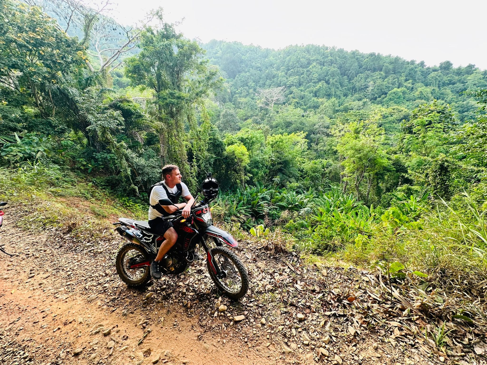 A man sitting on a dirt bike on a trail in a lush green forested area with mountains in the background.