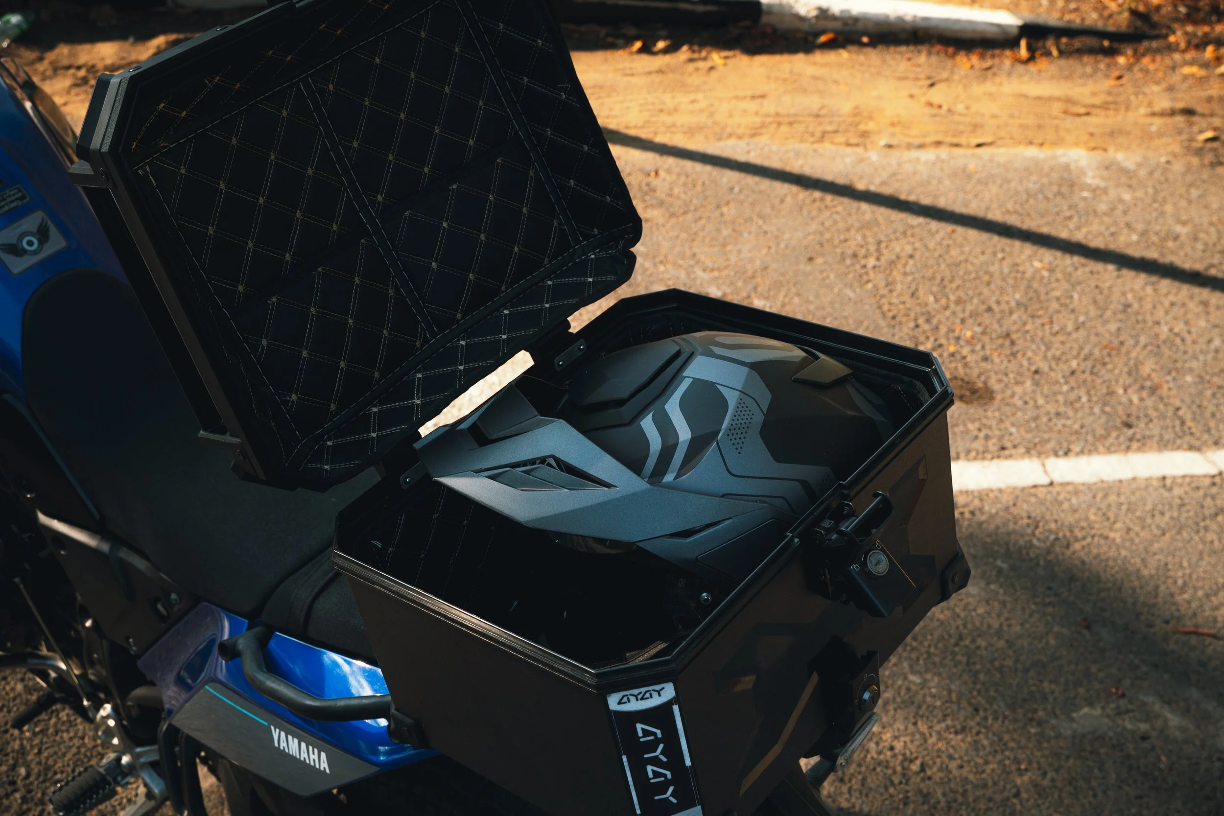 A motorcycle with a helmet stored in an open storage compartment on the back, parked on asphalt ground with some shadows.