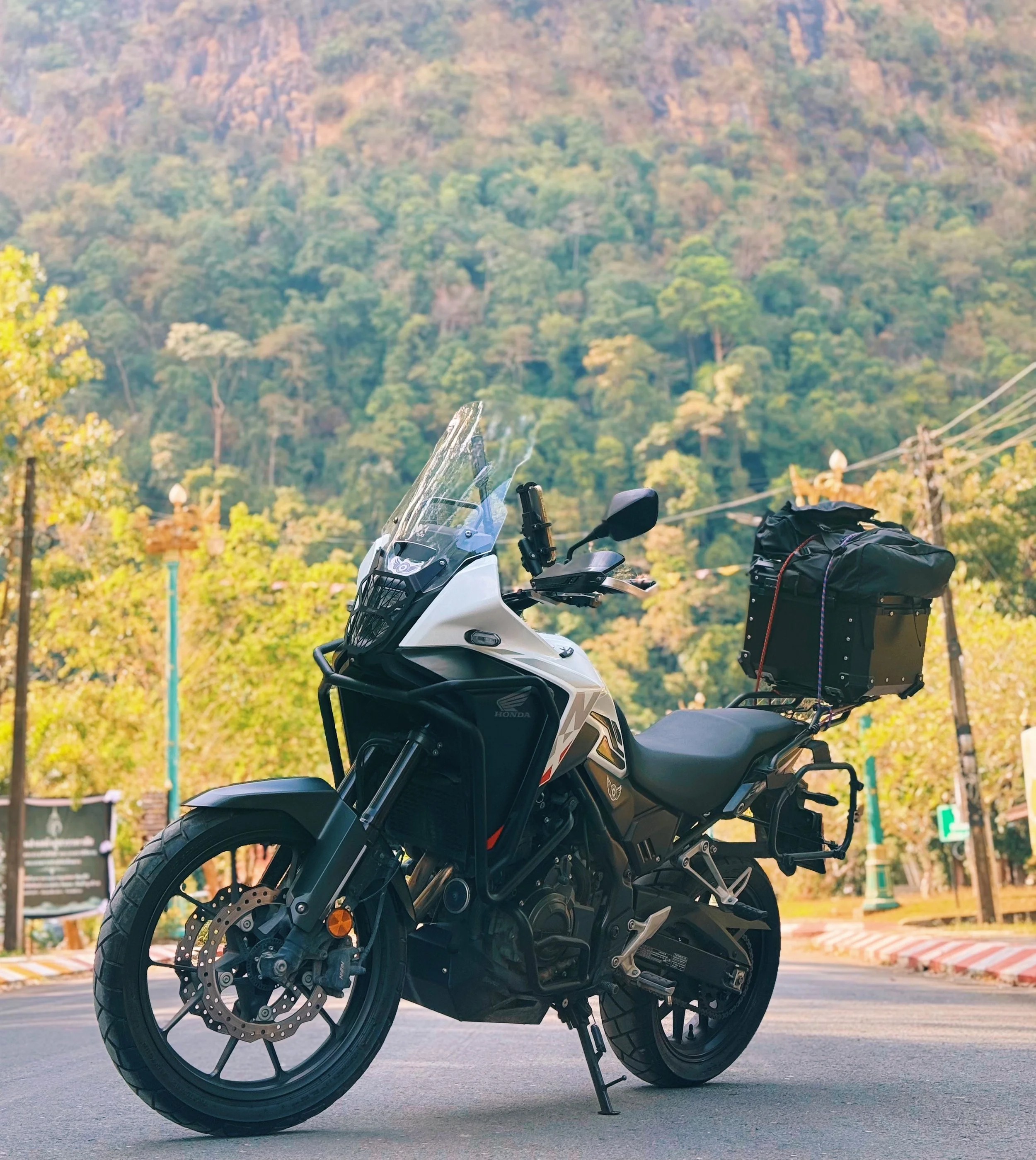 A black and white Honda motorcycle parked on a street with a green wooded hillside and mountain in the background.
