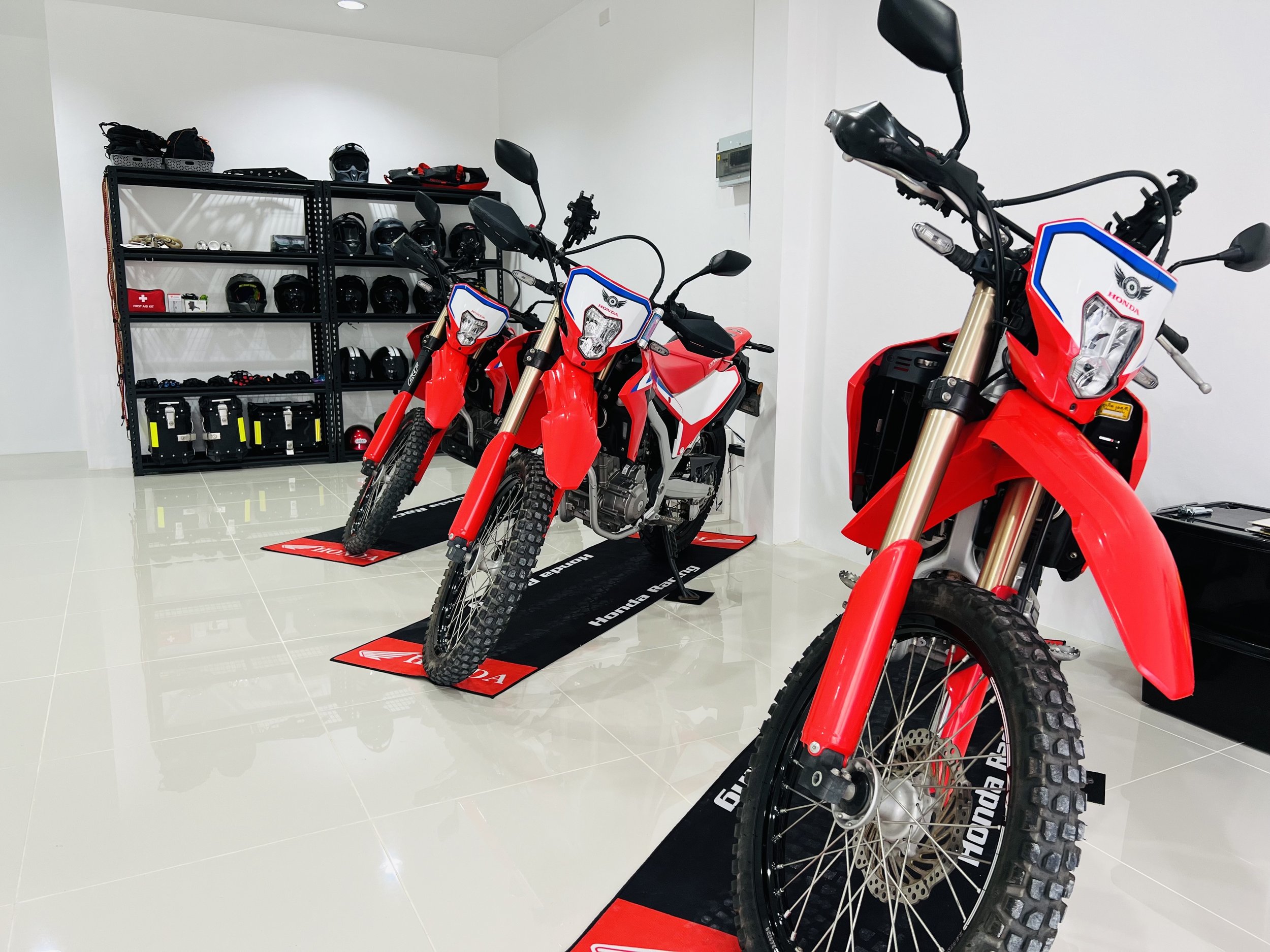 Three red Honda off-road motorcycles positioned on Honda branded mats inside a showroom. A black shelving unit in the background holds helmets and motorcycle gear.