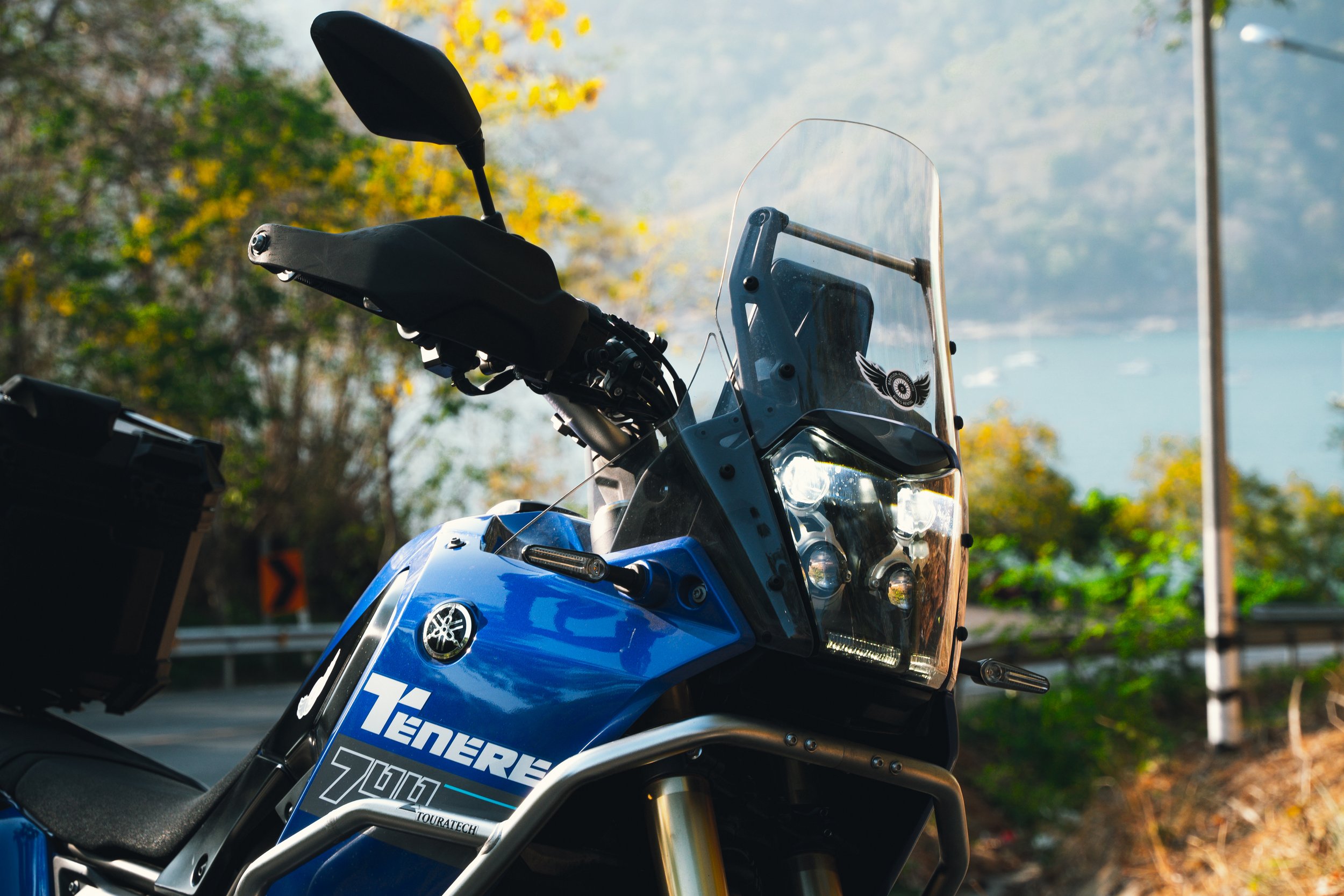 Close-up of a blue Yamaha Teneré Z201 motorcycle with a windshield and black handlebar, parked outdoors with trees and a body of water in the background.