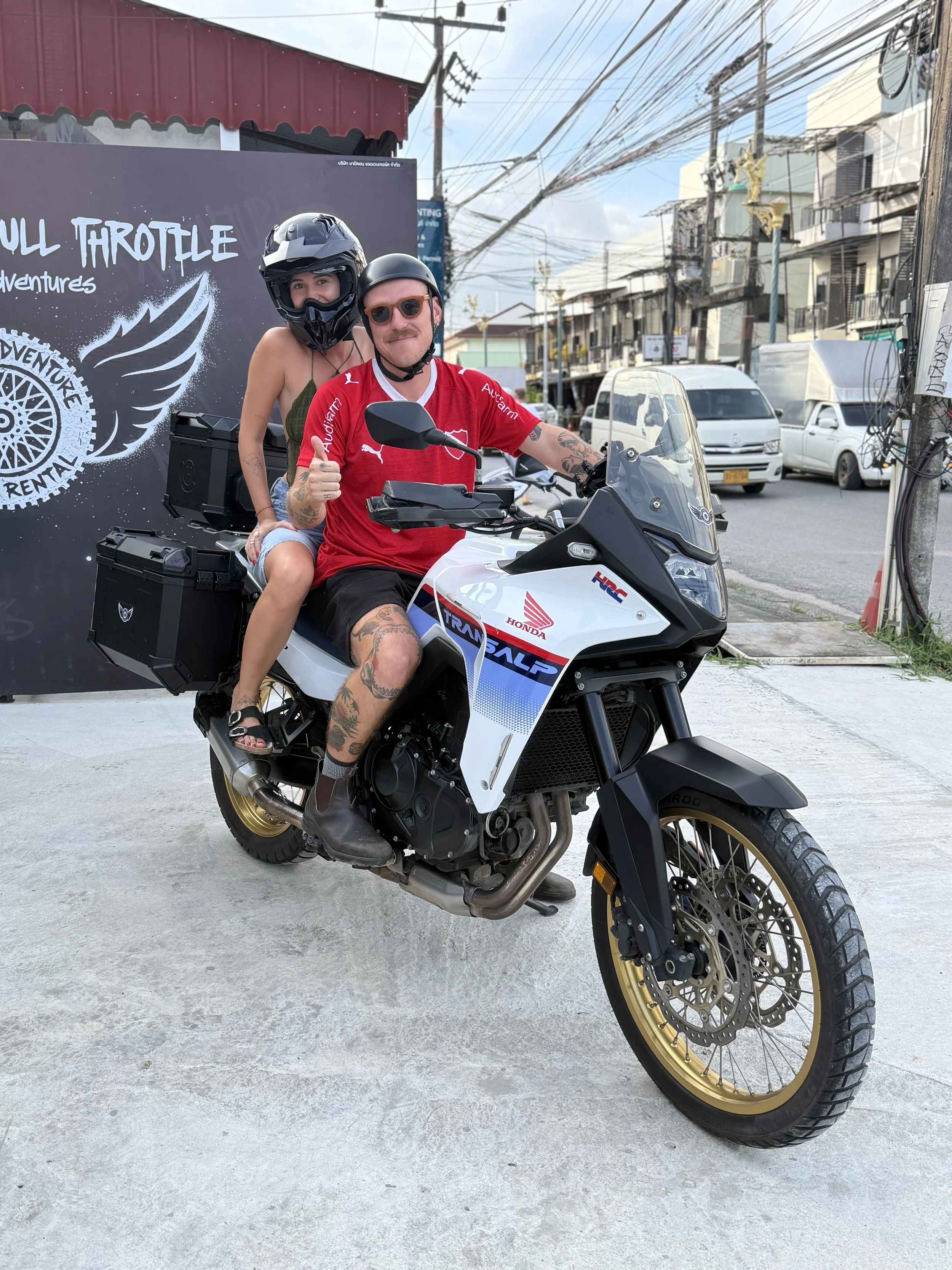 Two people sitting on a white Honda Transalp motorcycle on a street, with one person giving a thumbs-up. Both are wearing helmets, and the background features a sign and power lines.