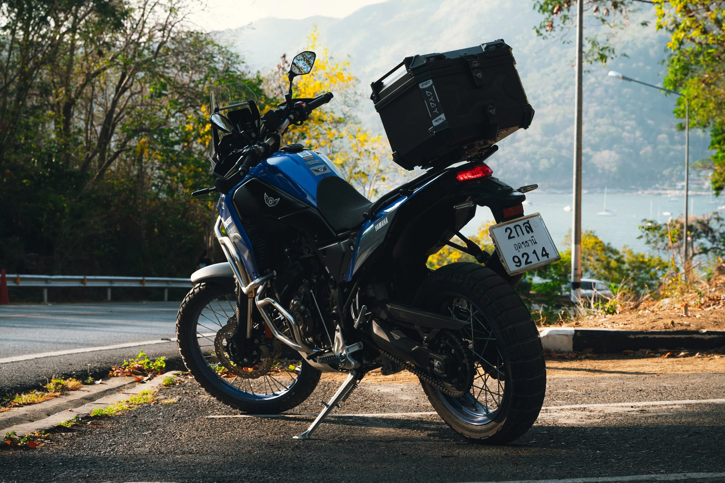 A blue and black motorcycle parked on the roadside with a large black storage box on the back, surrounded by trees and a view of a body of water and mountains in the background.