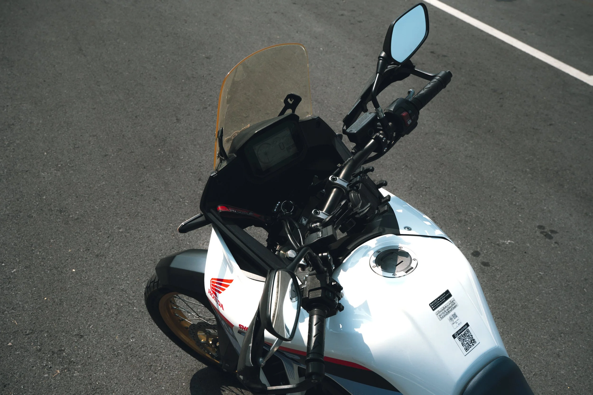 Top view of a white Honda motorcycle parked on asphalt, showing the handlebar, rearview mirror, transparent windscreen, and part of the fuel tank with a QR code sticker.