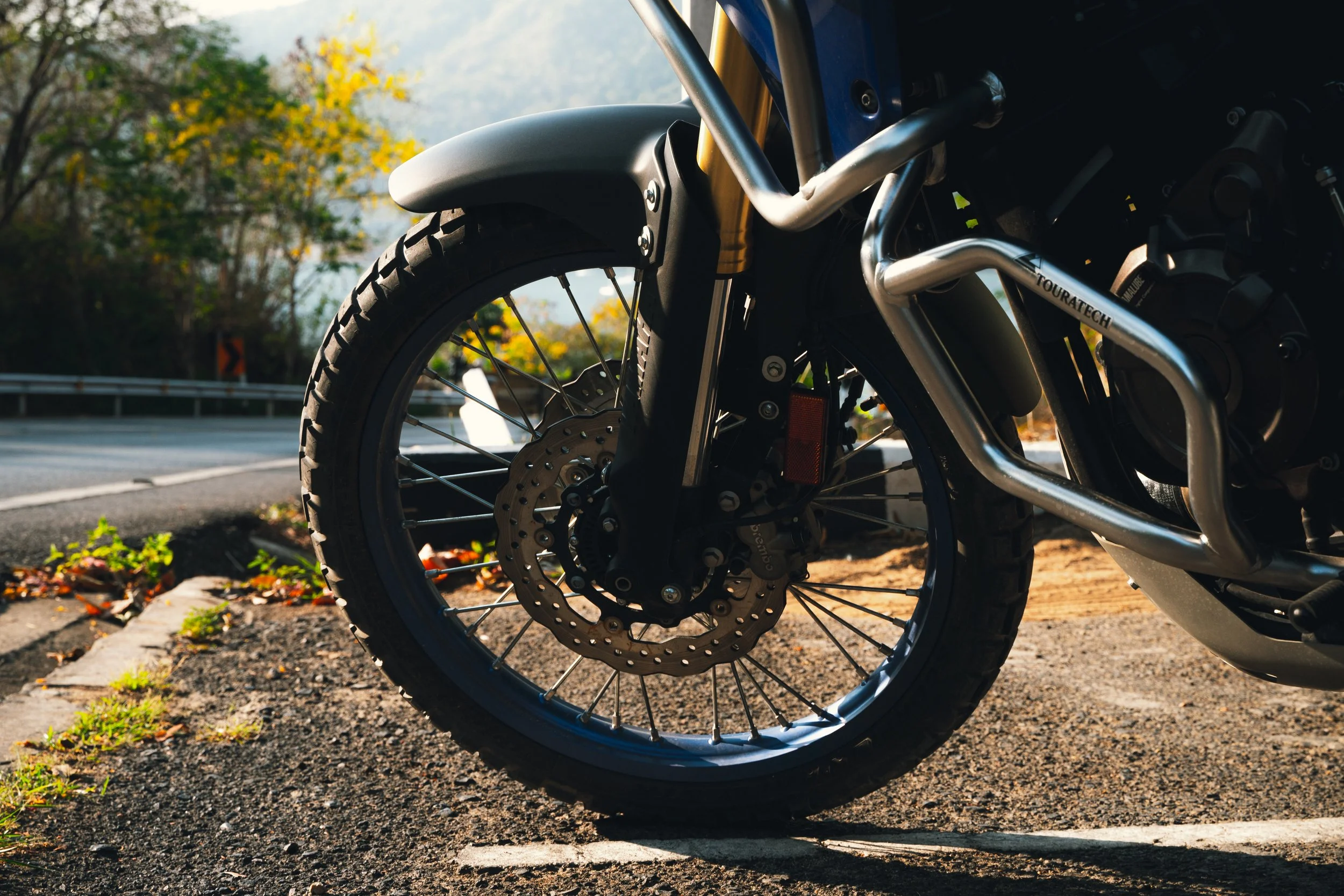 Close-up of a motorcycle wheel and front suspension on the side of a road with autumn foliage.