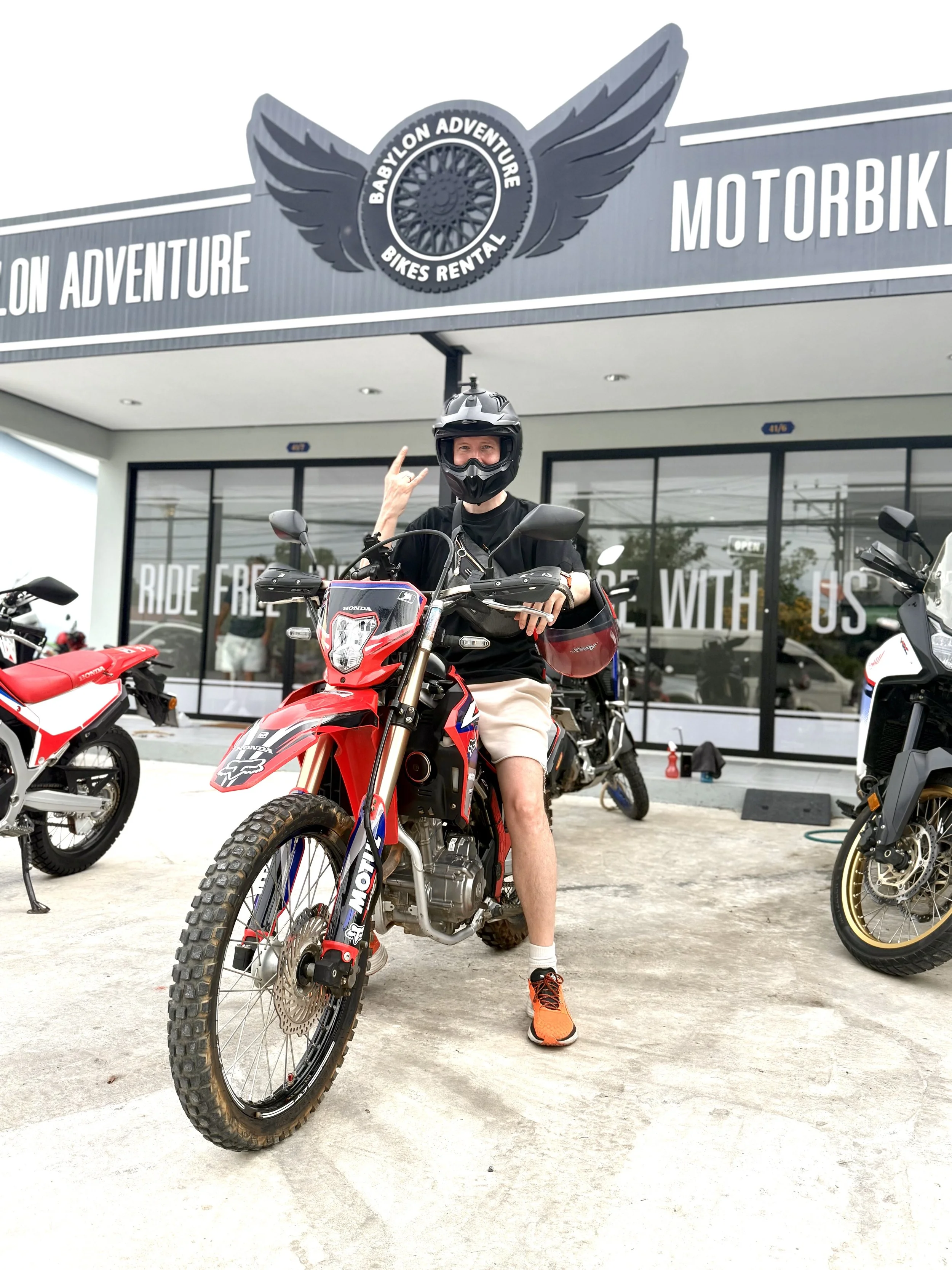 Person wearing black helmet and black shirt sitting on a red dirt bike outside motorcycle rental shop, other bikes parked nearby.