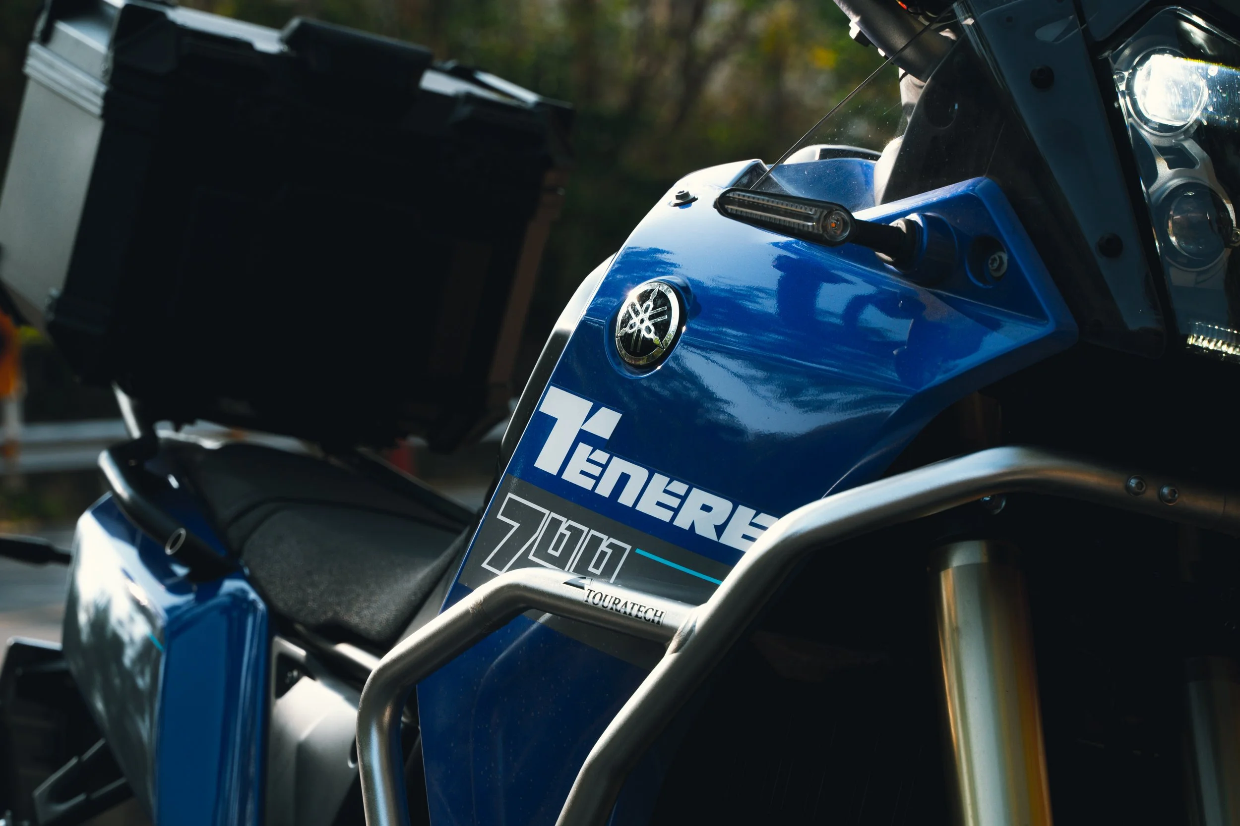Close-up of a blue Yamaha Tenere 700 adventure motorcycle with a top case mounted behind the seat, showing the Yamaha logo and Tenere 700 graphics on the side panel, with a partially visible headlight and front suspension.