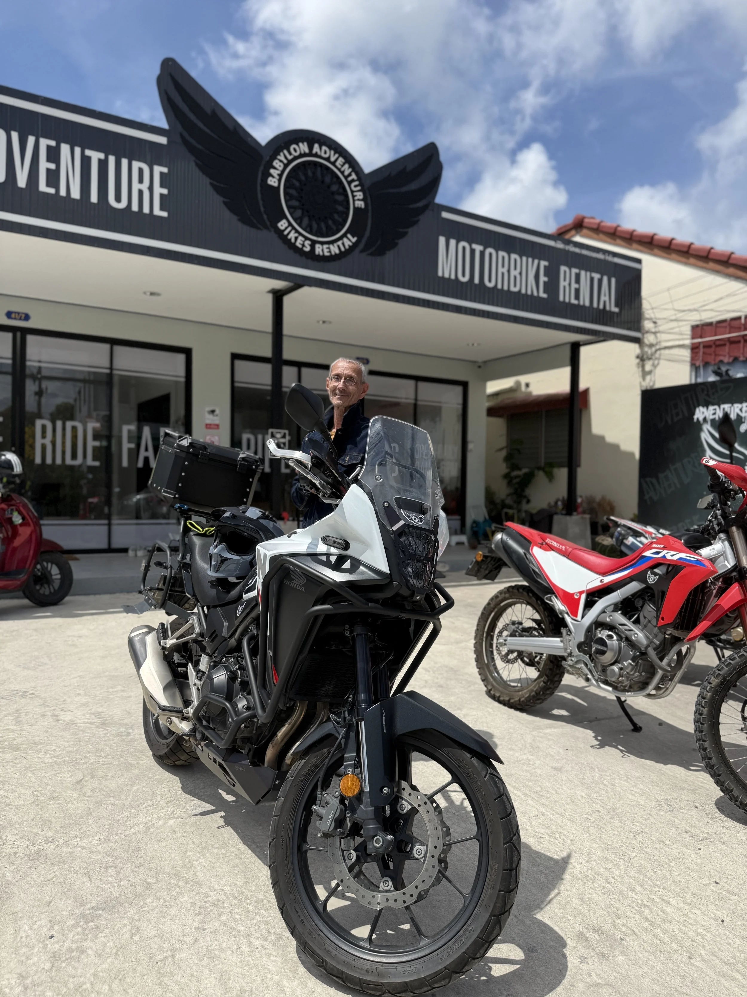 Man standing next to a black and white adventure motorcycle in front of a motorcycle rental shop with a sign reading Babylon Adventure Bikes Rental. Other motorcycles are parked nearby.