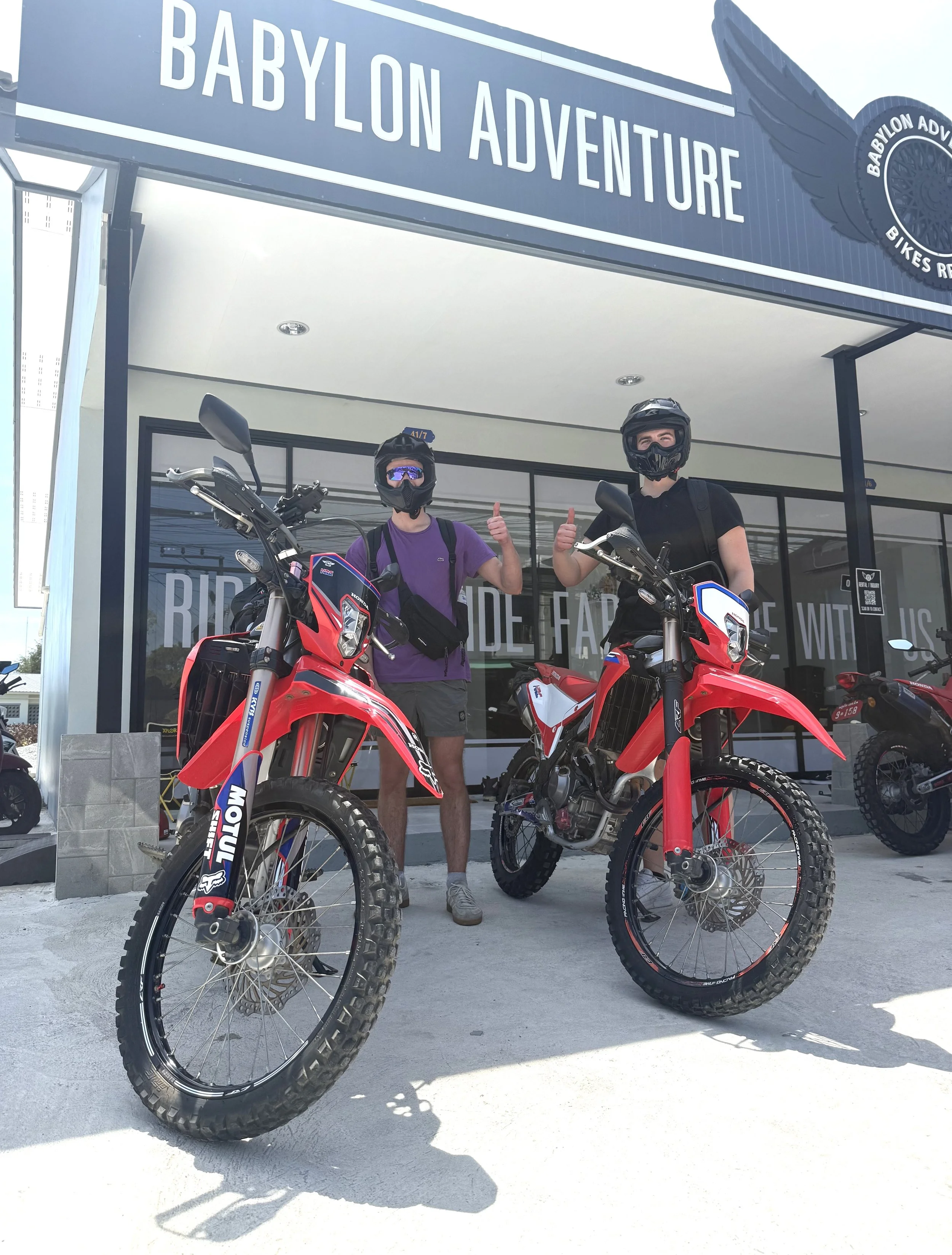 Two motorcyclists wearing helmets and protective gear standing with red off-road motorcycles in front of a building labeled 'Babylon Adventure'.