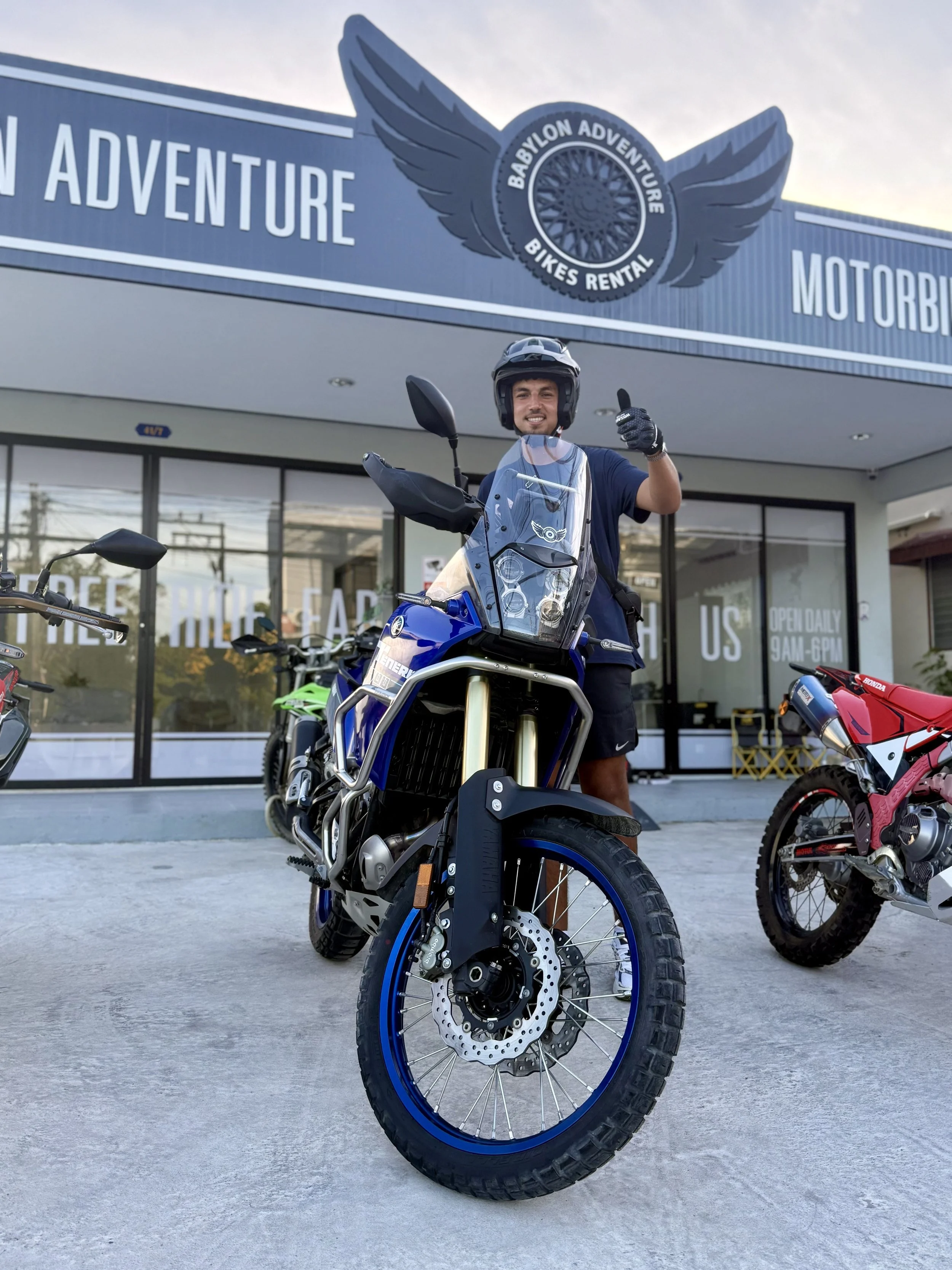 A man standing next to a blue Yamaha motorcycle in front of Babylon Adventure Bikes Rental shop, giving a thumbs-up while wearing a helmet and gloves. There are other motorbikes parked nearby, and the shop has large glass windows with signage.