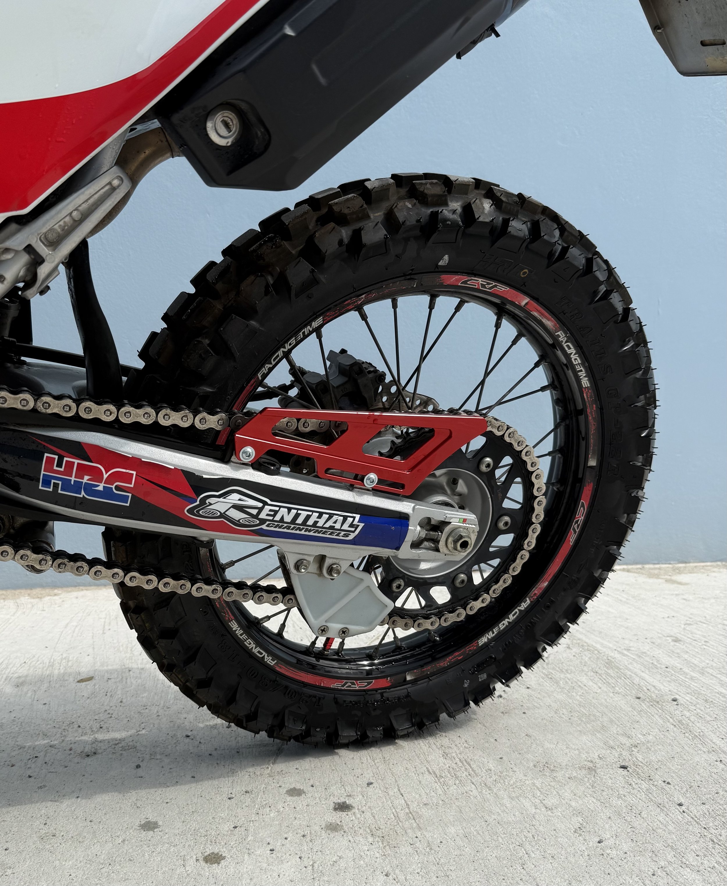 Close-up of a dirt bike rear wheel, tire, chain, and suspension, set against a light blue wall and concrete ground.