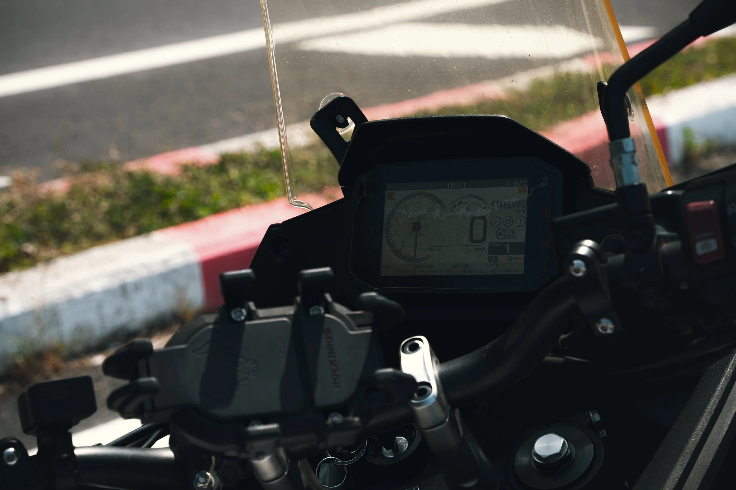 View of a motorcycle dashboard with a digital display, handlebar controls, and a windshield, with a curb and road in the background.