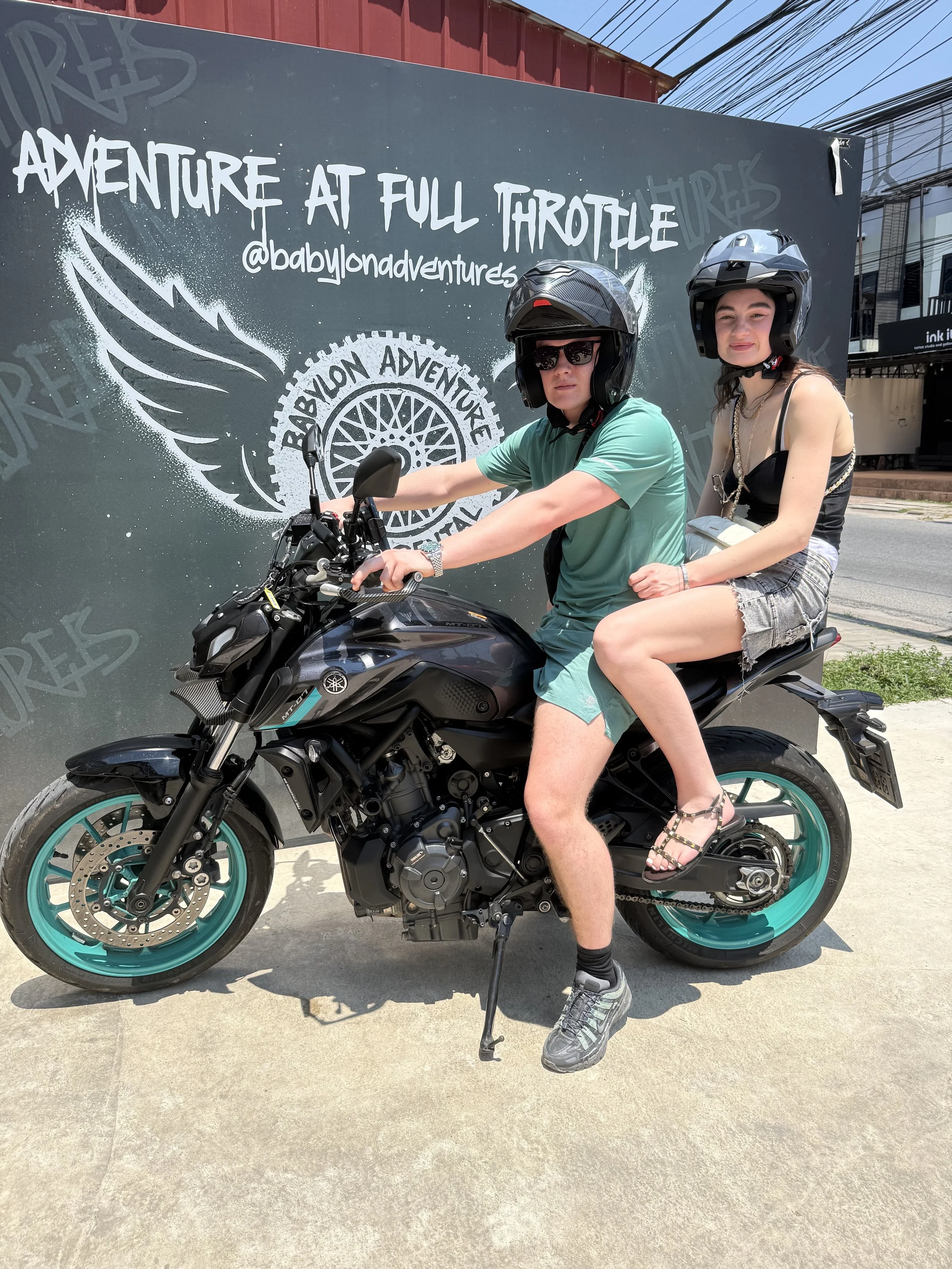 Two people wearing motorcycle helmets riding a black and teal motorcycle in front of a black wall with white text and a graphic design. The wall has the words 'Adventure at full throttle' and a social media handle '@babylonadventures.'