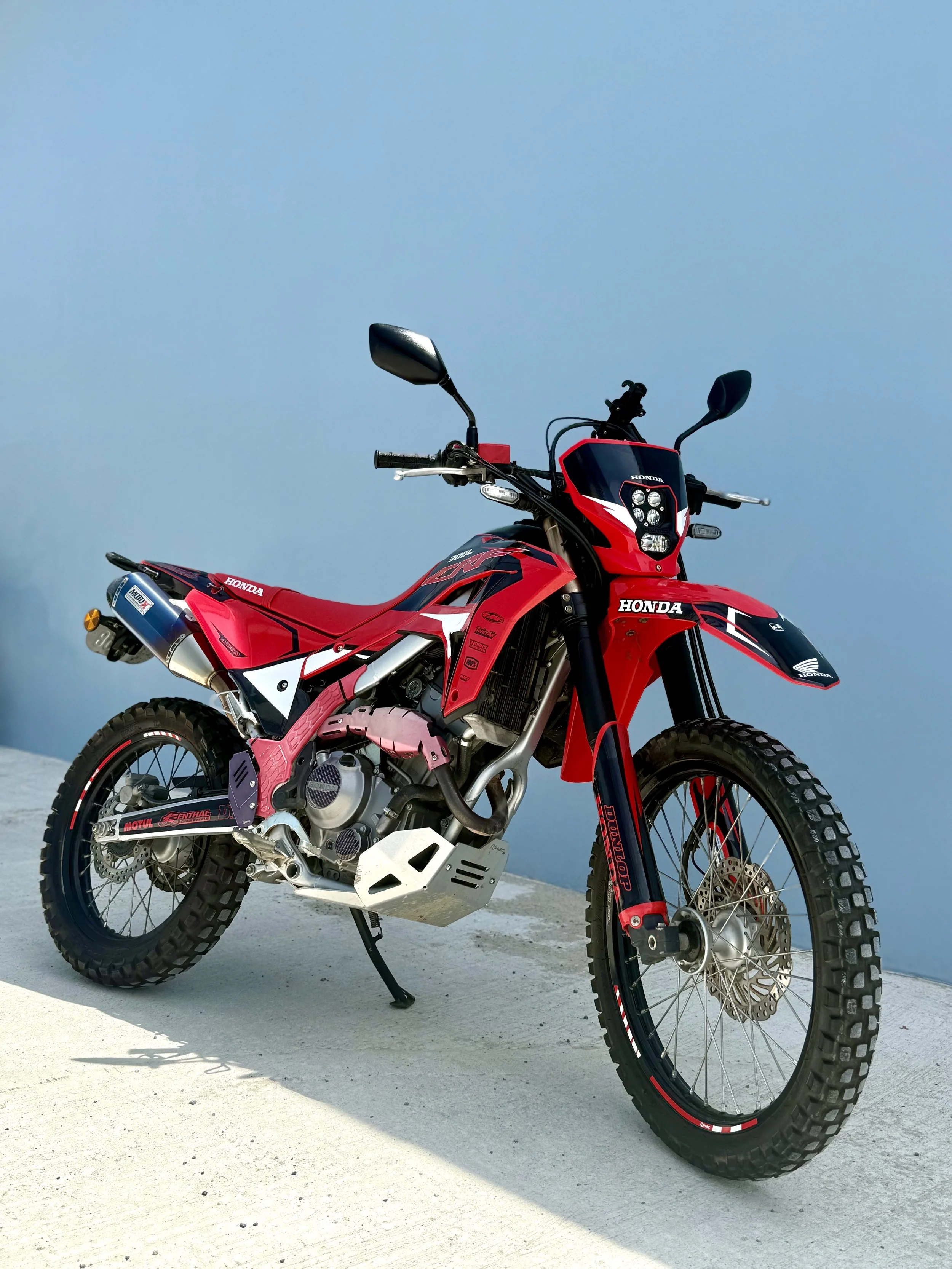 Red and black Honda off-road motorcycle on a light-colored surface with a blue sky background.