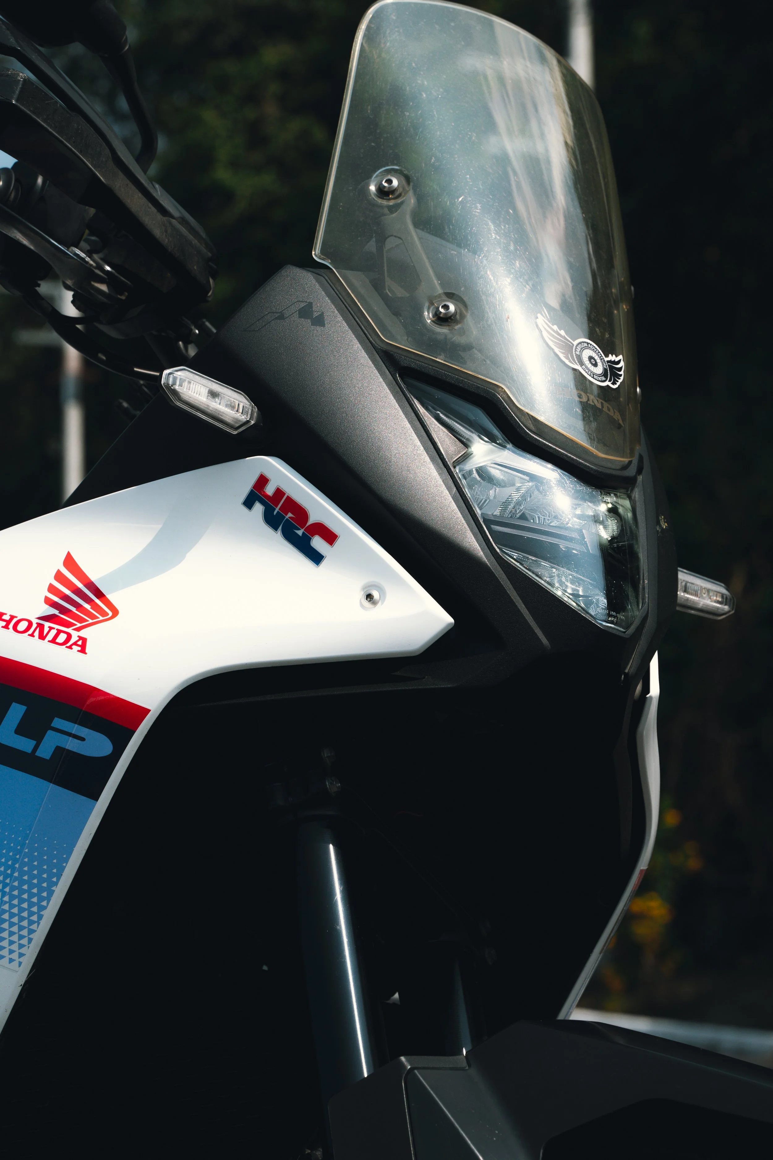 Close-up of a Honda Africa Twin motorcycle, showing the front with stickers including HRC, with a transparent windshield and green and trees in the background.