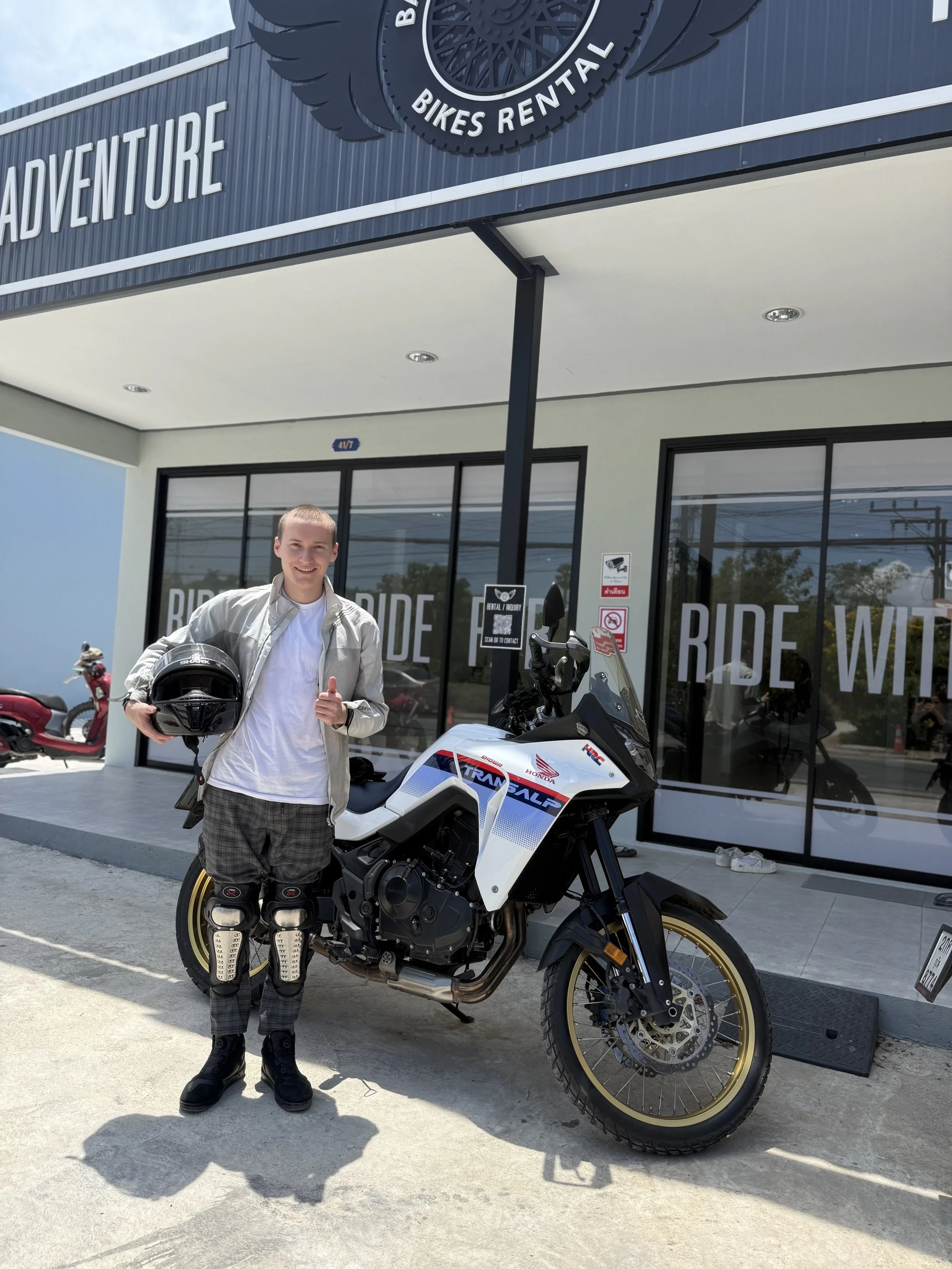 Young man standing outside a motorcycle rental shop, holding a helmet and giving a thumbs up next to a white Honda Transalp adventure motorcycle.