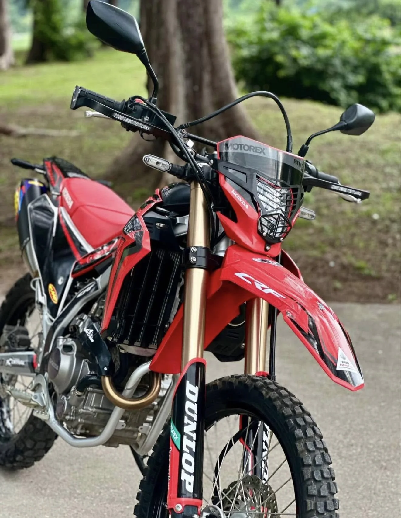 Red Honda off-road motorcycle with DUNLOP tires, black and white accents, parked outdoors on a paved path with green trees in the background.