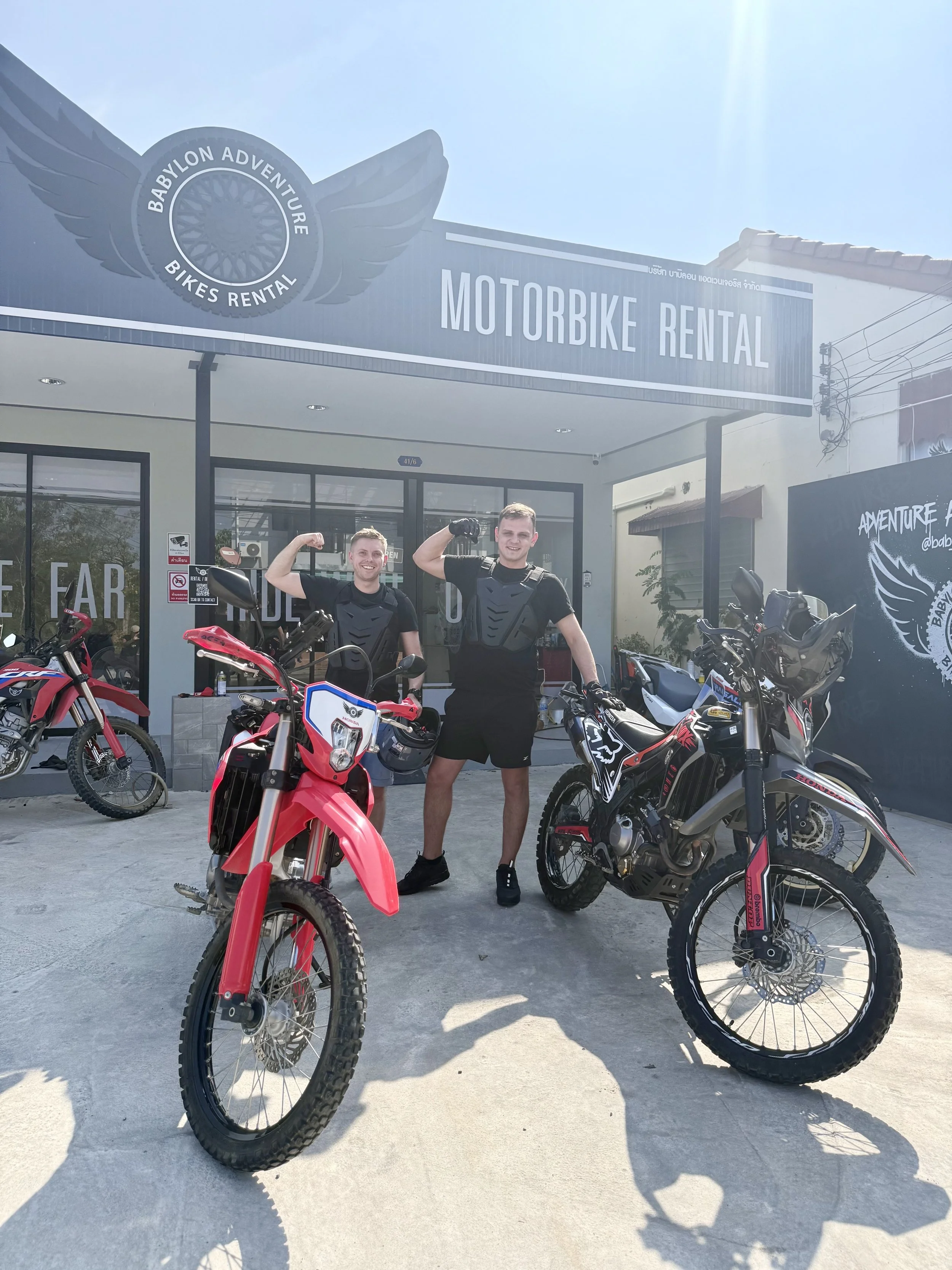 Two men in motorcycle gear standing in front of a motorcycle rental shop with off-road motorcycles, smiling and posing for the photo.