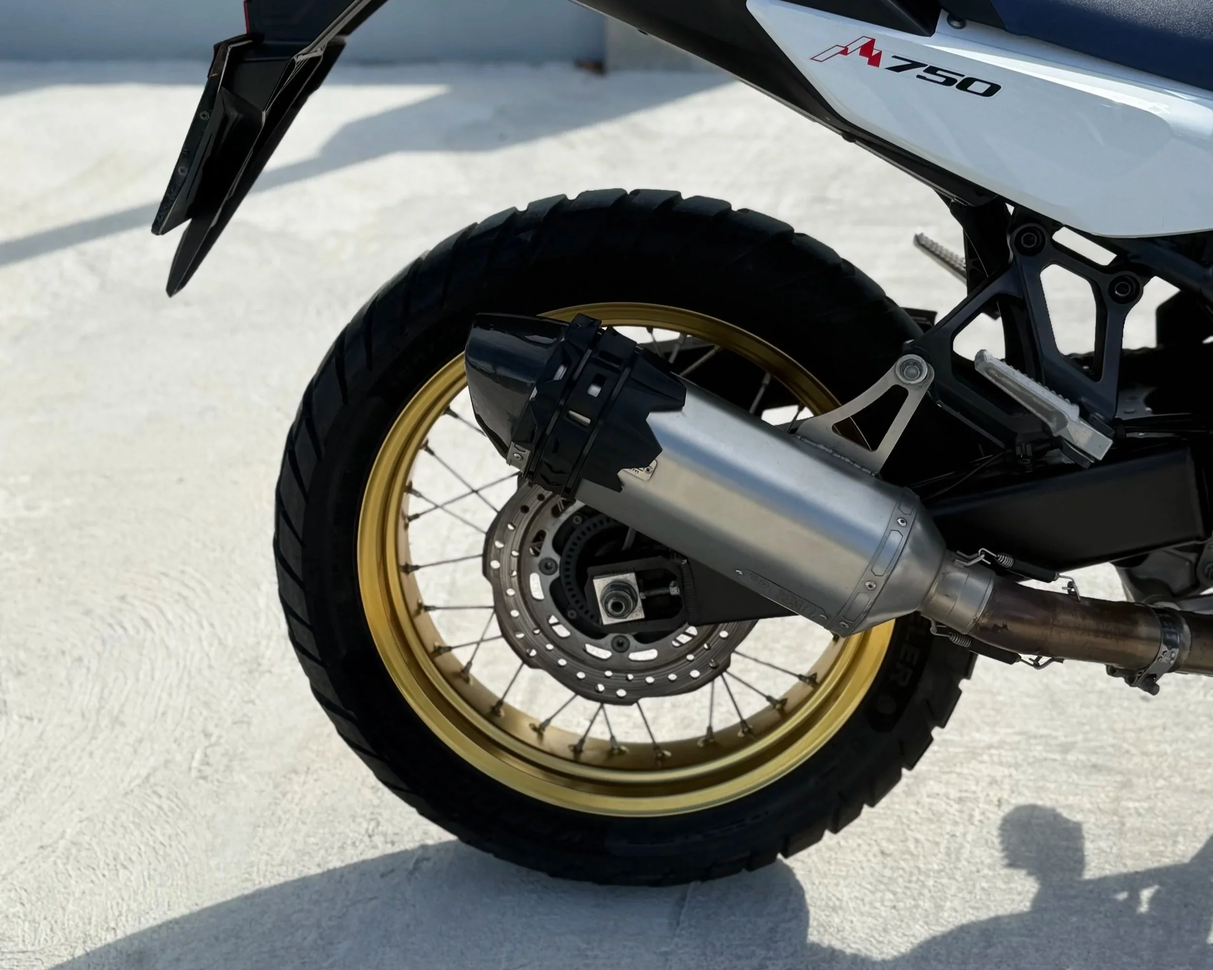 Close-up of a motorcycle's rear wheel, showing a gold rim, disc brake, exhaust pipe, and a part of the motorcycle frame with the label 'M750'.