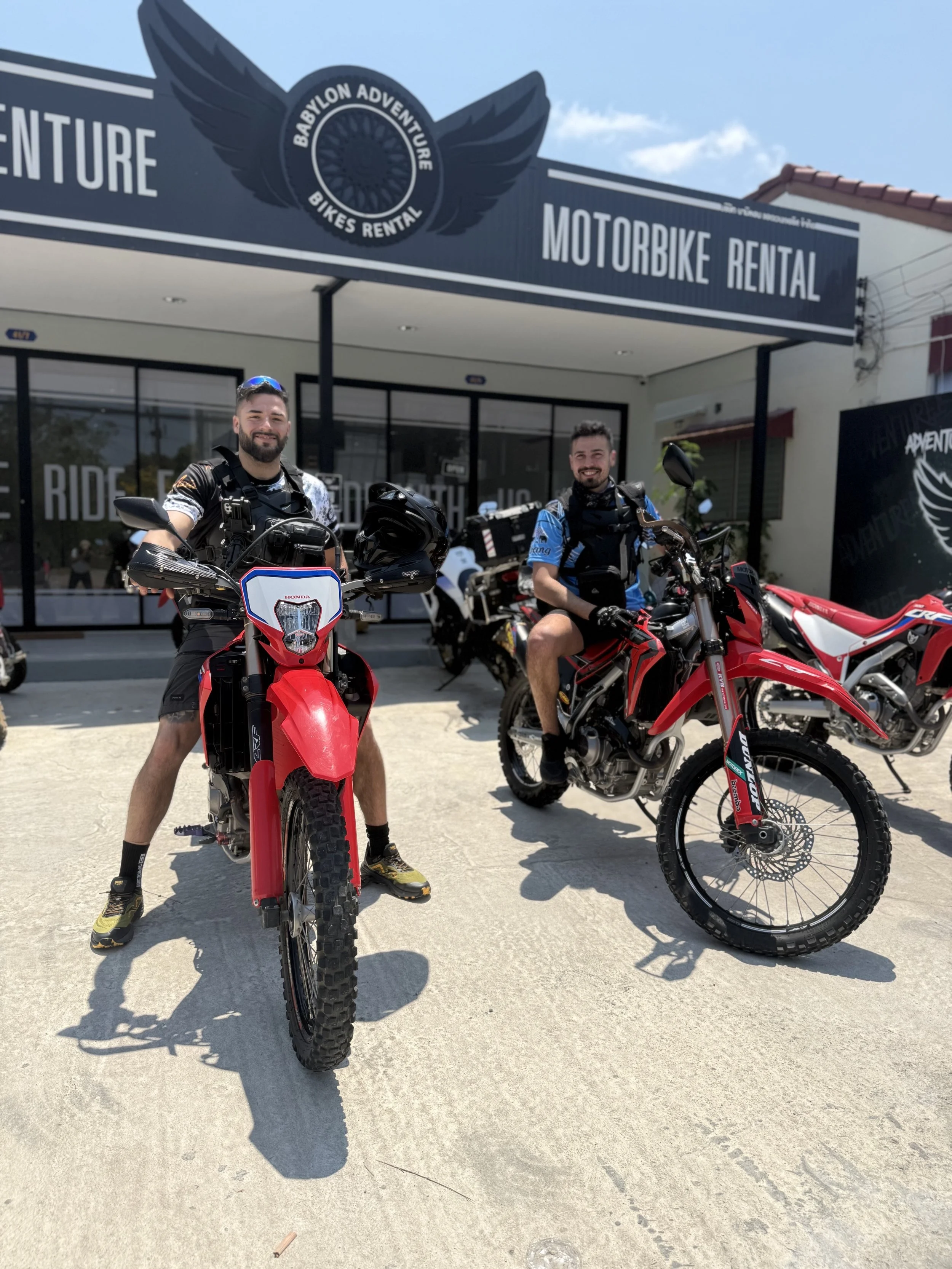 Two men in riding gear sitting on red dirt bikes in front of a motorcycle rental shop with a sign that reads 'Babylon Adventure Bikes Rental.'