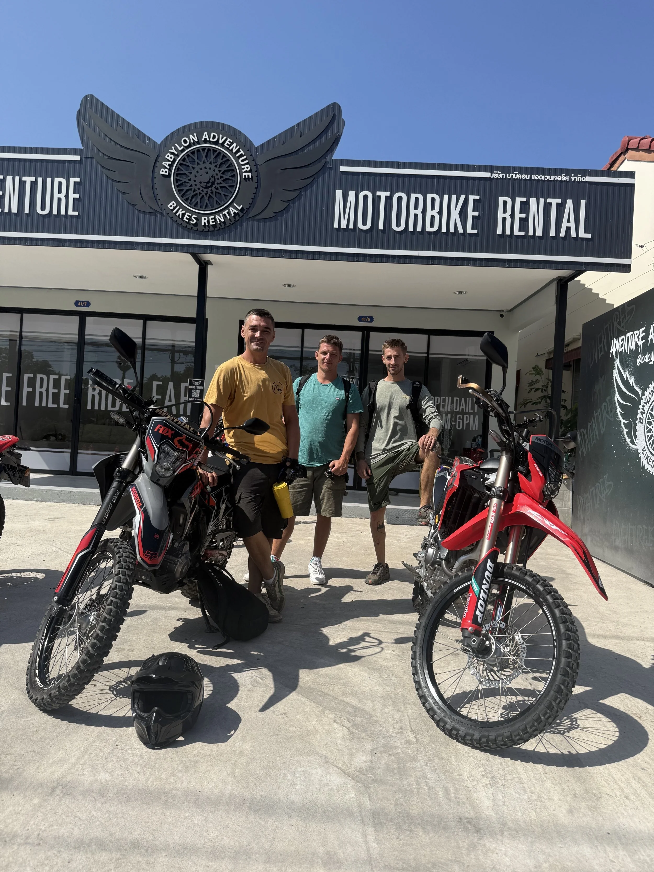 Three men standing in front of a motorcycle rental shop with two off-road motorcycles, a helmet on the ground, and a blue sky overhead.