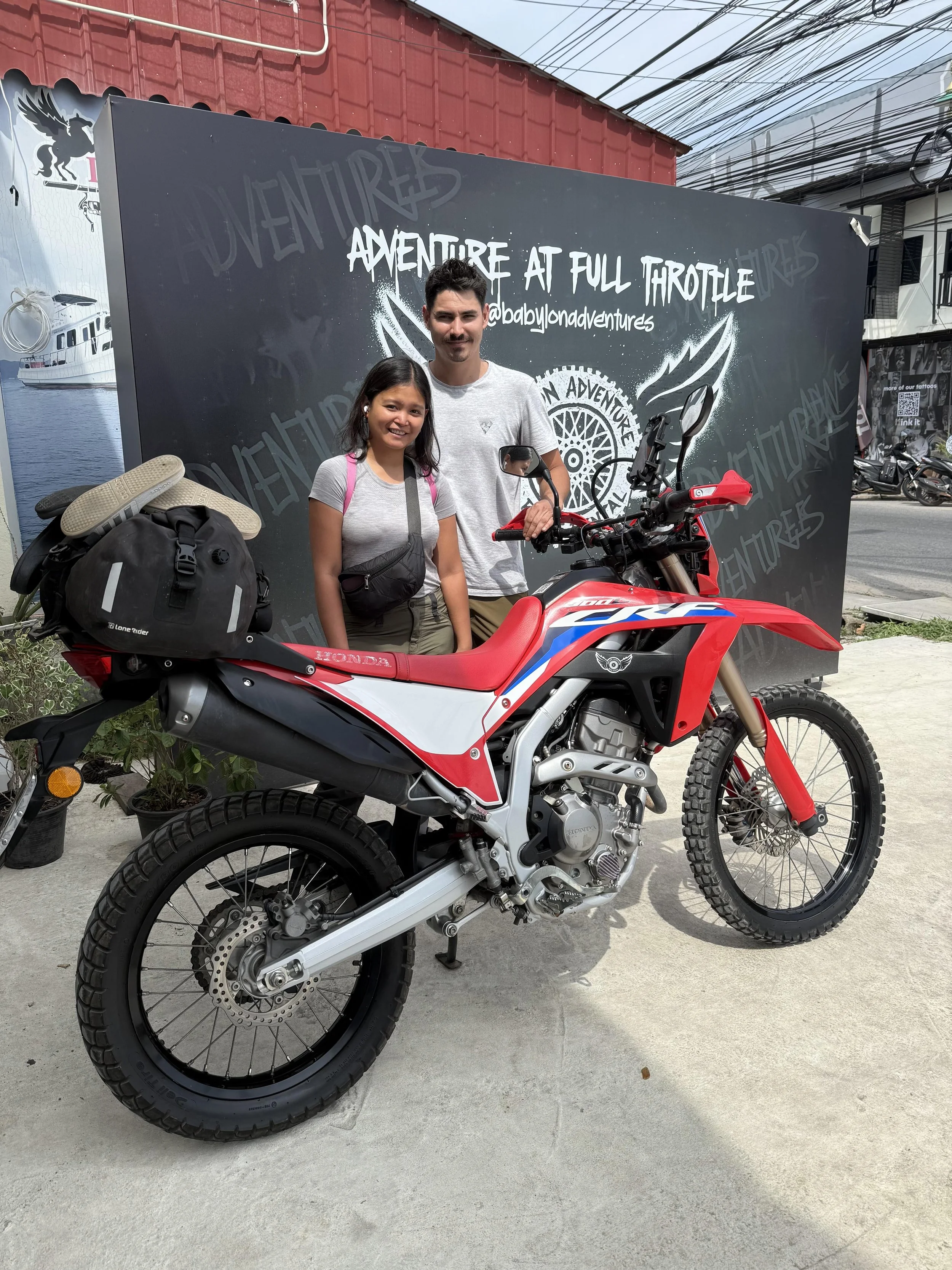 A young woman and a young man standing next to a red and black off-road motorcycle in front of a black sign that reads 'Adventure at Full Throttle' and '@babylondonadventures'. The woman is smiling, carrying a pink backpack and has a black waist bag,