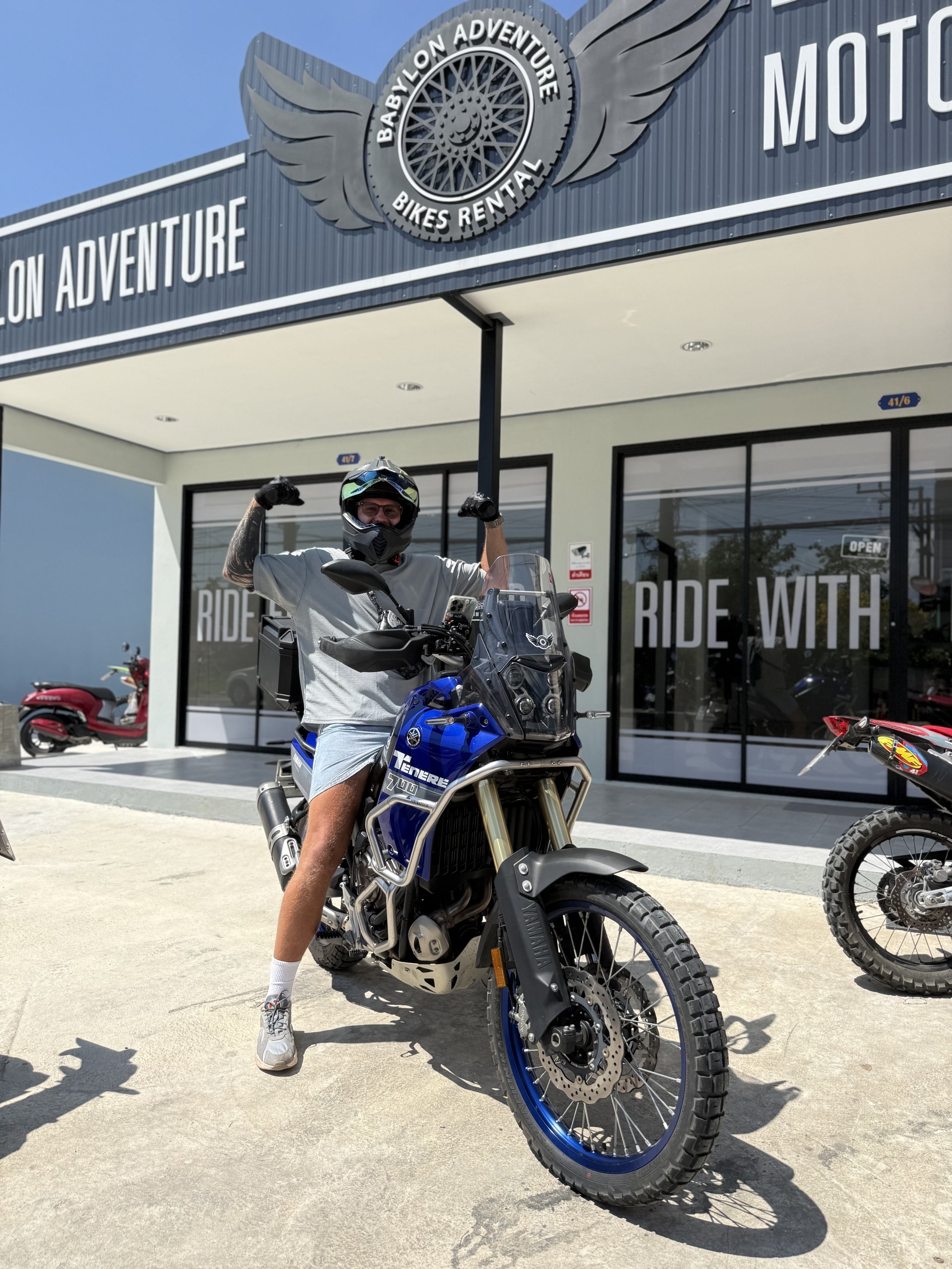 A person wearing a helmet and sunglasses sitting on a Yamaha Ténéré 700 motorcycle in front of Babylon Adventure bike rental shop, flexing their arms. The shop has a sign that says "Ride With" and other motos parked nearby.