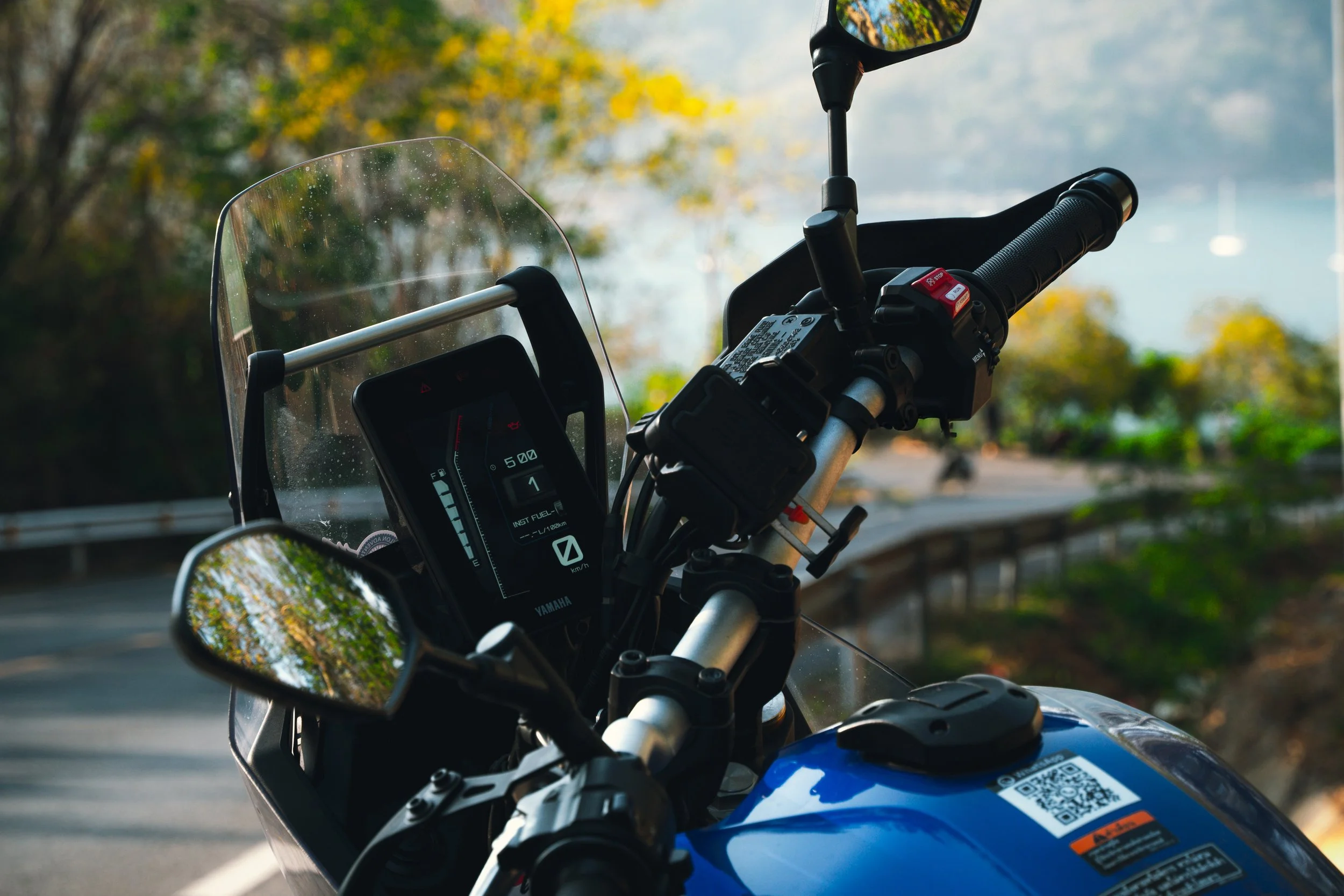 Close-up of a blue motorcycle with a digital dashboard, side mirror, handlebar, and a QR code sticker on the fuel tank, set against a scenic outdoor background with trees and a guardrail.