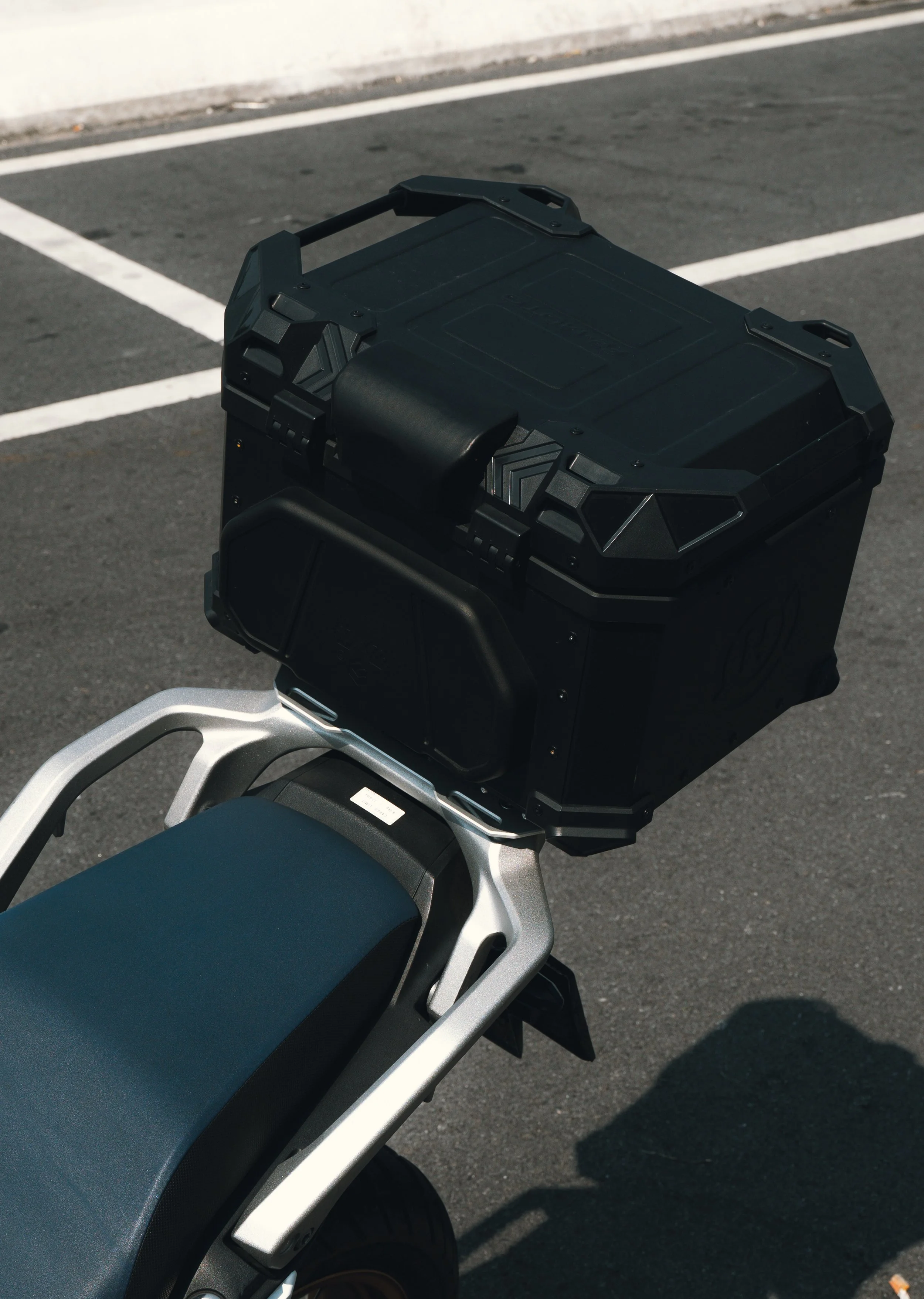 Close-up of a motorcycle seat and a black storage box mounted on the rear, parked on an asphalt surface with white parking lines.