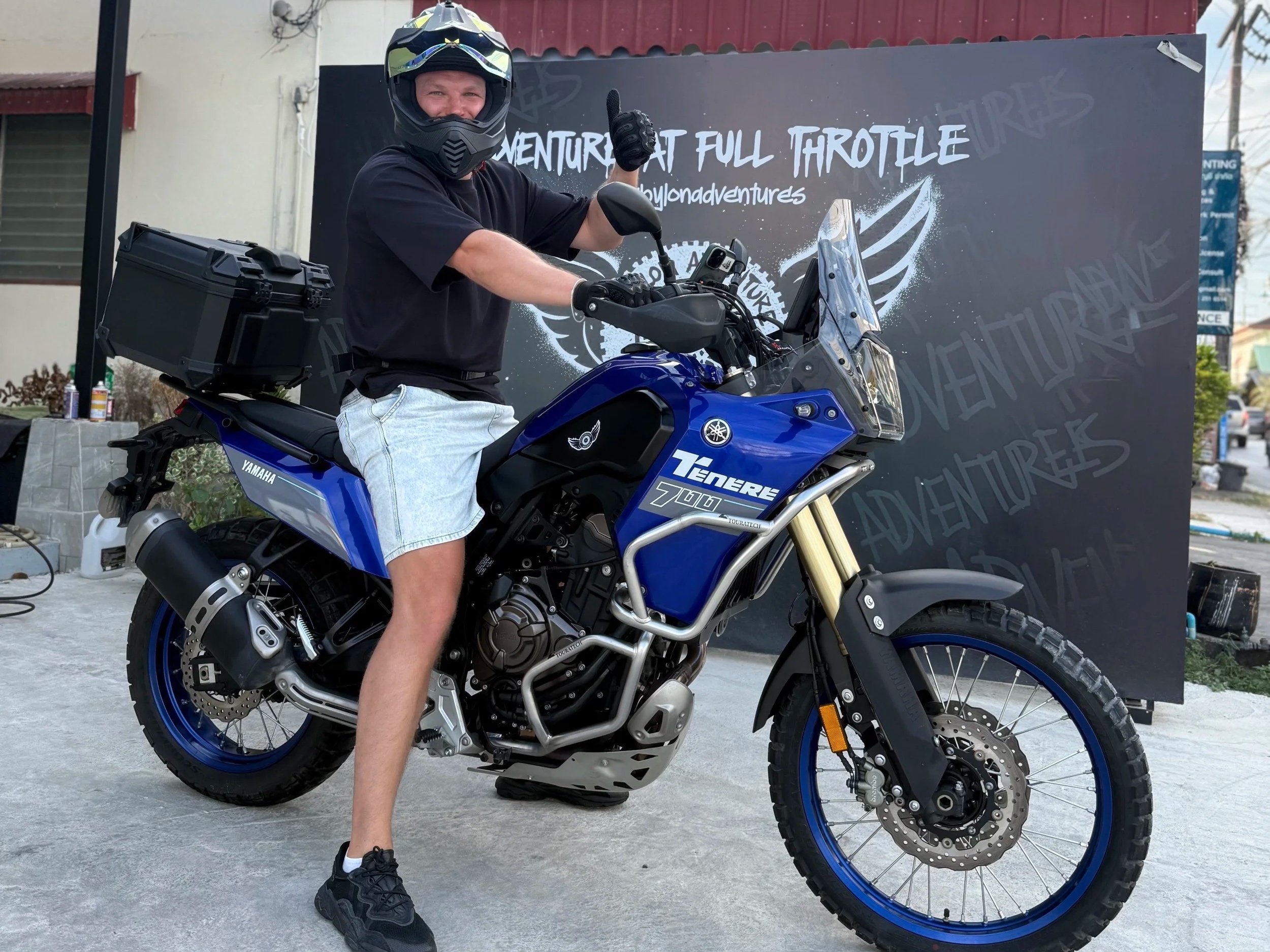A man wearing a black helmet and black t-shirt giving a thumbs-up while sitting on a blue Yamaha Teneré 700 motorcycle, parked outdoors with a black backdrop that reads 'Adventure at Full Throttle'.