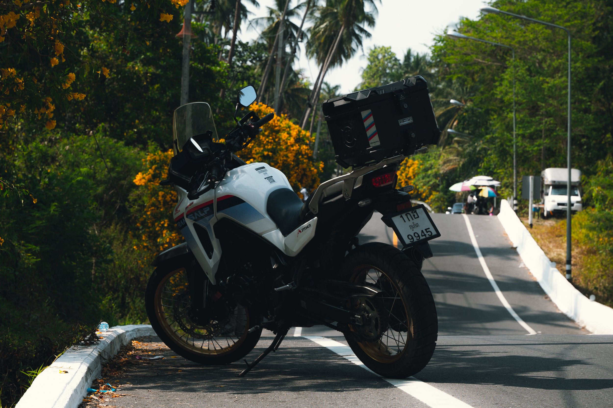 A white off-road motorcycle parked on the side of a scenic road with lush green trees and yellow flowering bushes, and a delivery box mounted on the back.