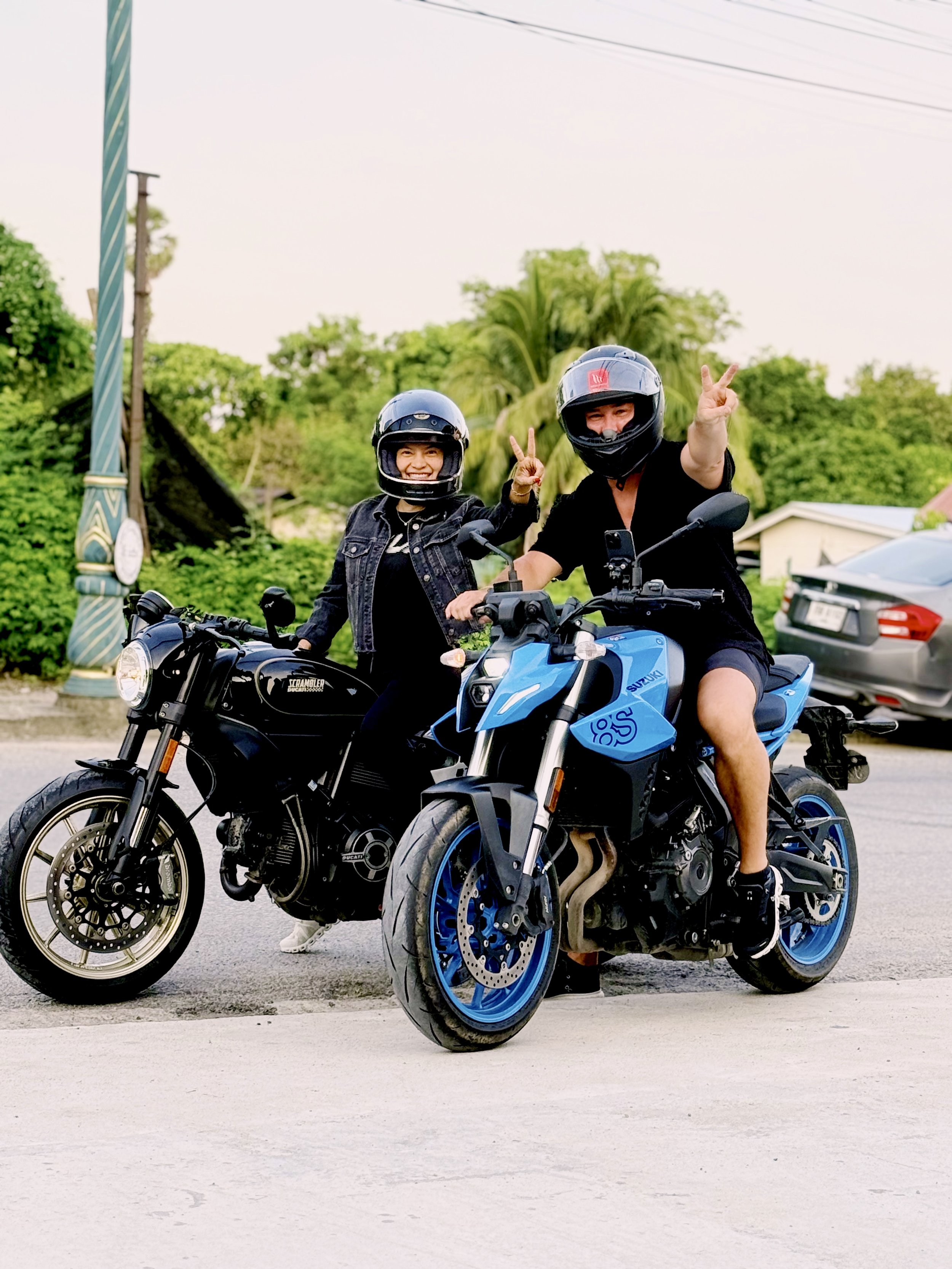 Two people wearing helmets on motorcycles, smiling and making peace signs.