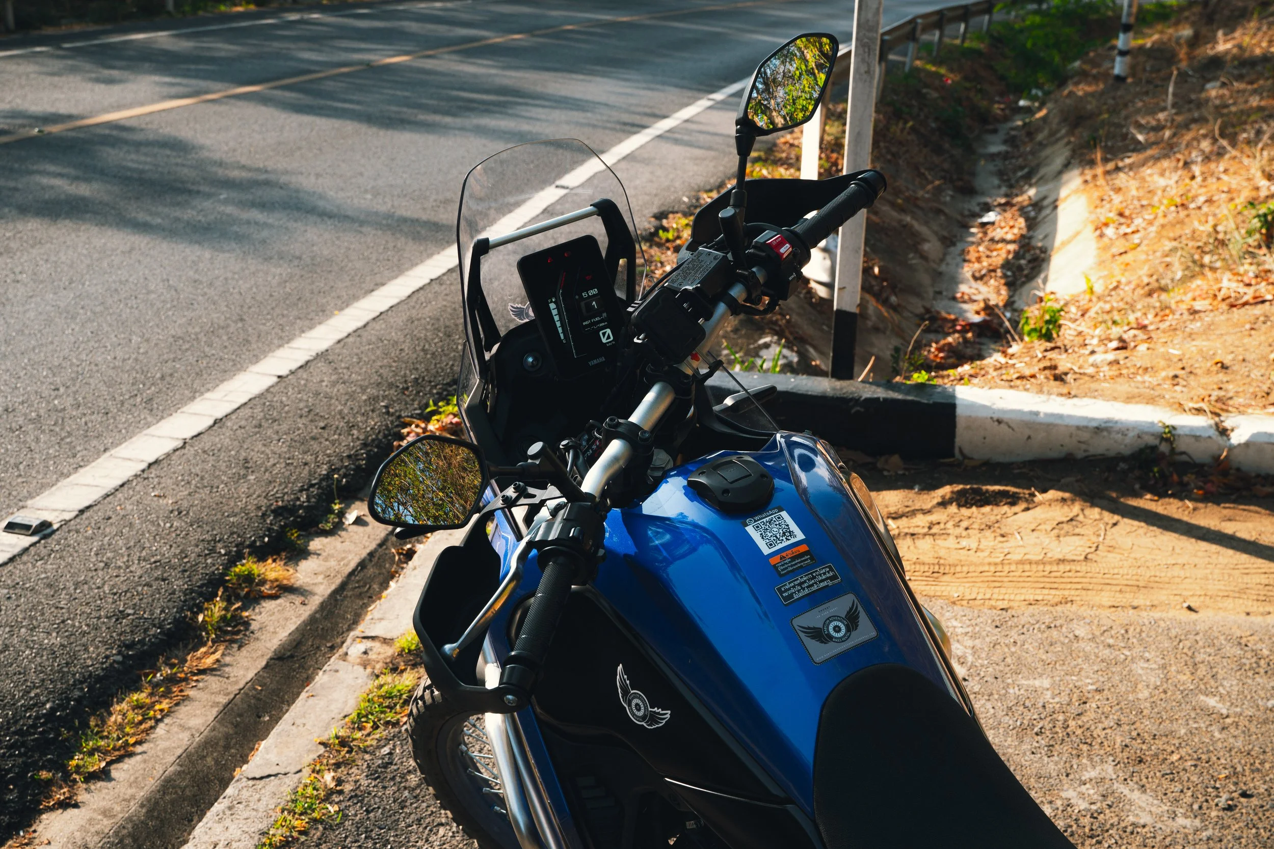 Blue motorcycle parked on the side of the road next to a curb and a guardrail, with trees reflected in its mirrors and shadow on the ground.