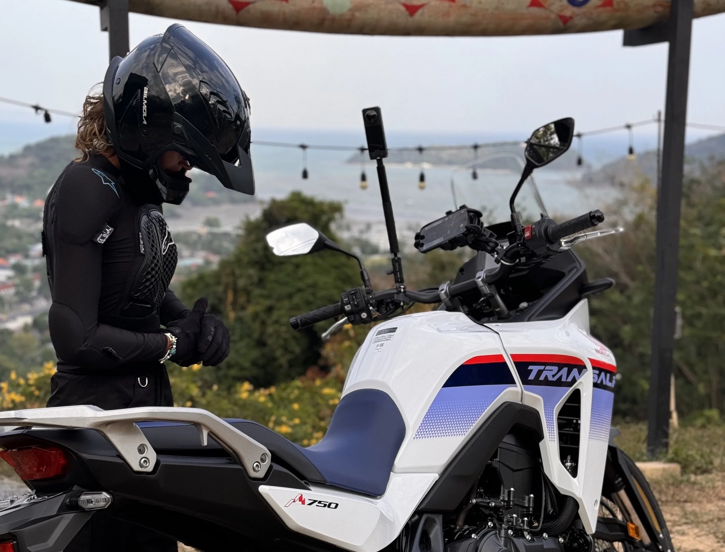 A person in a black helmet and riding gear preparing to ride a white motorcycle with blue, red, and black accents outdoors against a background of trees and distant hills.
