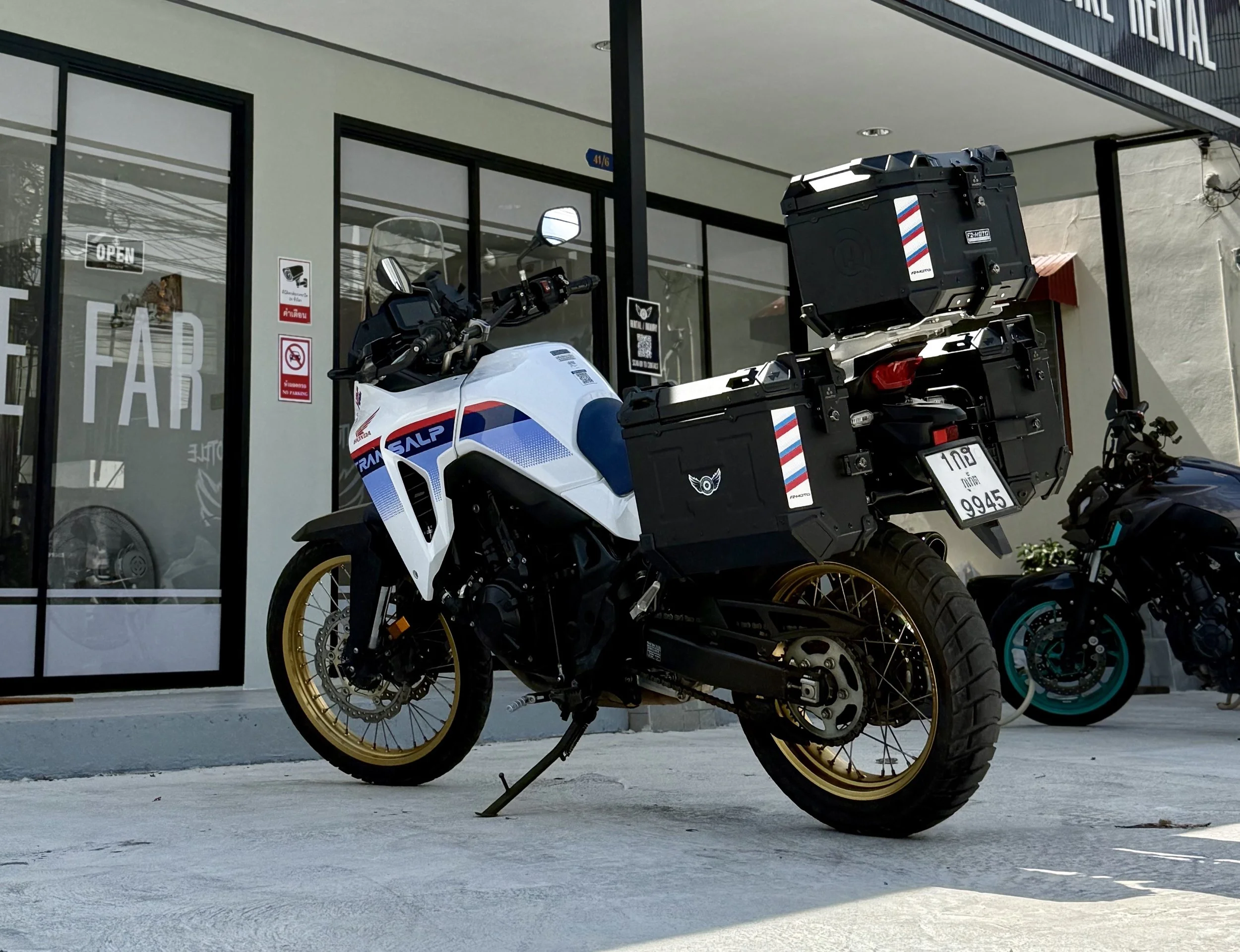 A white Honda motorcycle equipped with black and gold luggage cases parked outside of a building with glass doors and windows.
